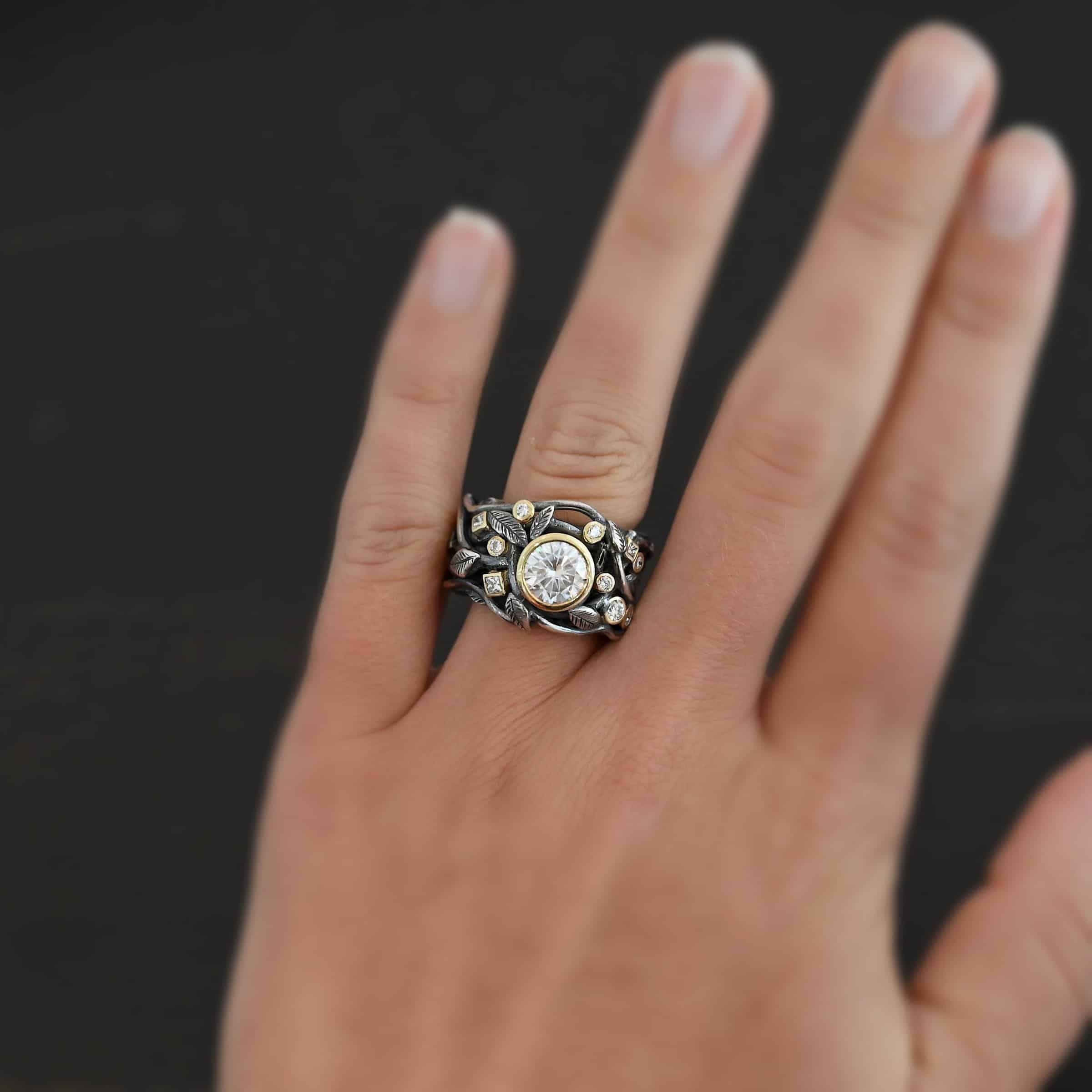 Enchanted Ring Photo of the "Enchanted" Ring. Ring has a large moissanite in the center and smaller moissanites scattered around the entire ring. There are multiple silver leaves around the entire ring. There are eleven stones total, the center stone, and ten moissanites around the rest of the ring, five on each side. Each stone is set in gold. Ring is worn on a models engagement ring finger to demonstrate size.