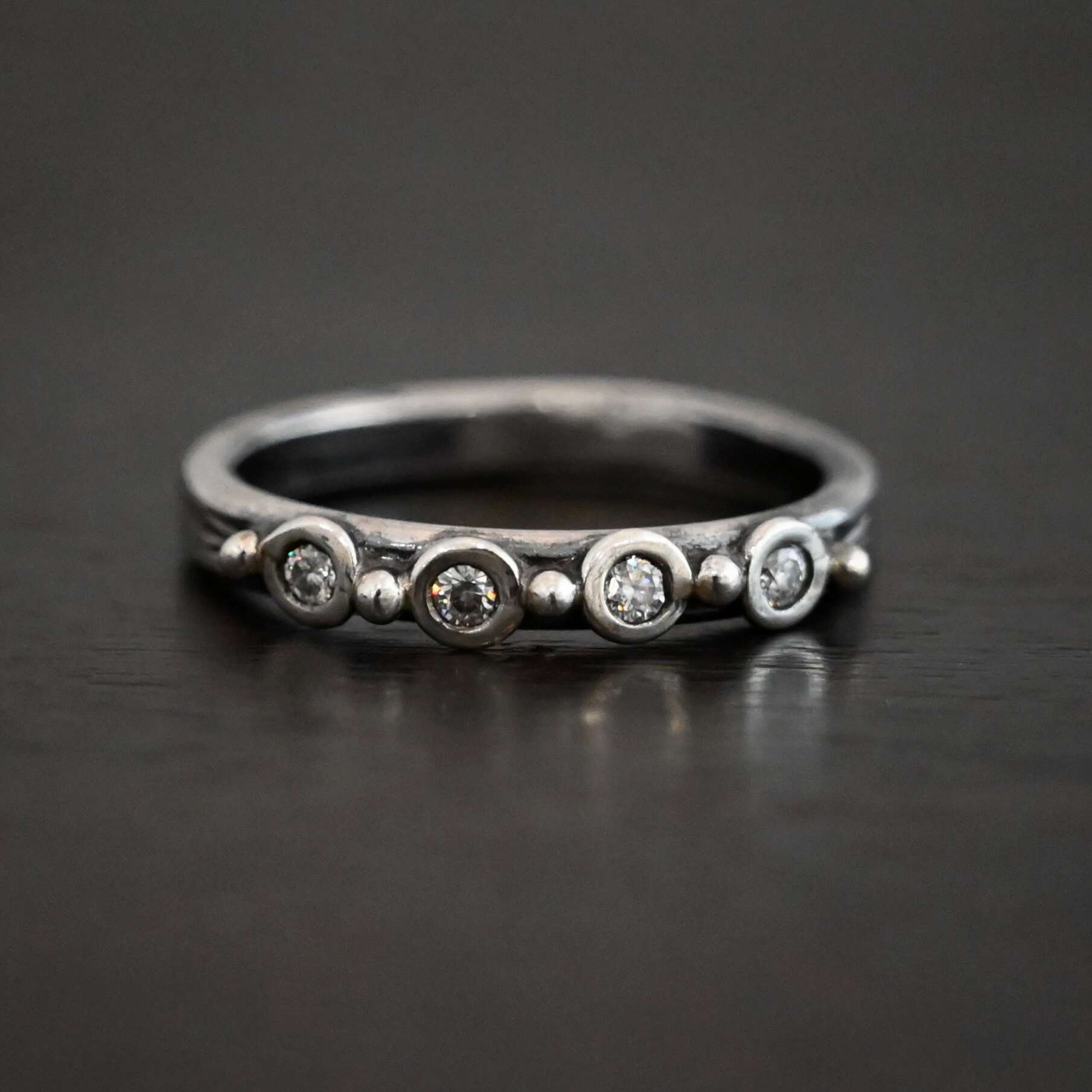 Photo of the "Years Of Love" Anniversary Band. It is a simple silver band with four diamonds across it. There are five silver balls on the band. Three are separating each diamond, and there is a silver ball before the first diamond and after the last diamond. Ring is sitting against a dark background.