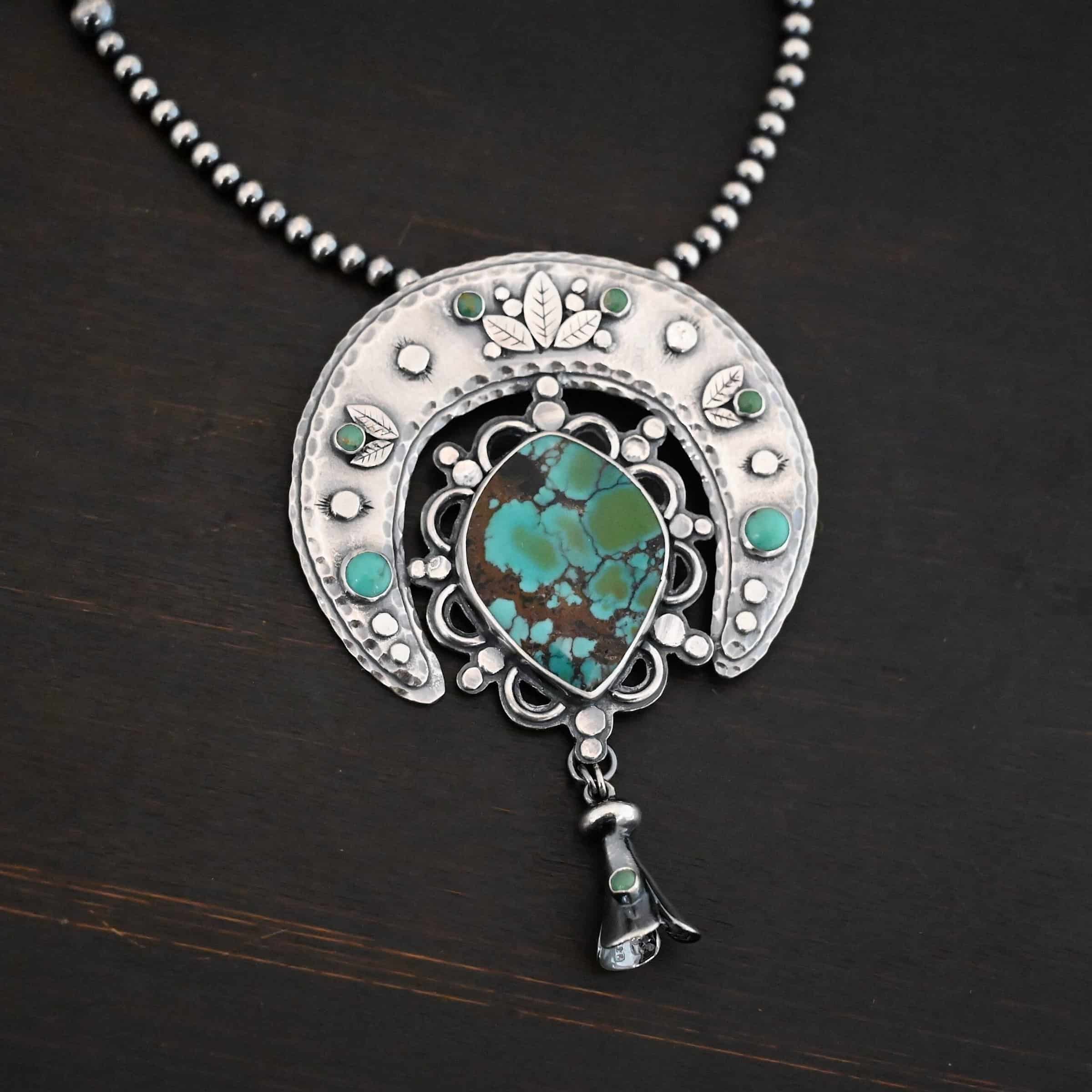 Wanderer Squash Blossom Necklace Photo of the "Wanderer Squash Blossom" Necklace. It has a large turquoise necklace on a black background. It is a squash blossom necklace. There is a large turquoise in the center with a flower petal design around it. There are seven other small turquoise on the horseshoe shaped silver piece going around the flower design of the turquoise. This piece has silver leaves and hammered silver dots on it as well. Hanging beneath the turquoise is a small cone of silver with cuts in it to make it look like an upside down flower petal. The chain is beaded and has the cone silver flower petals scattered around the chain.