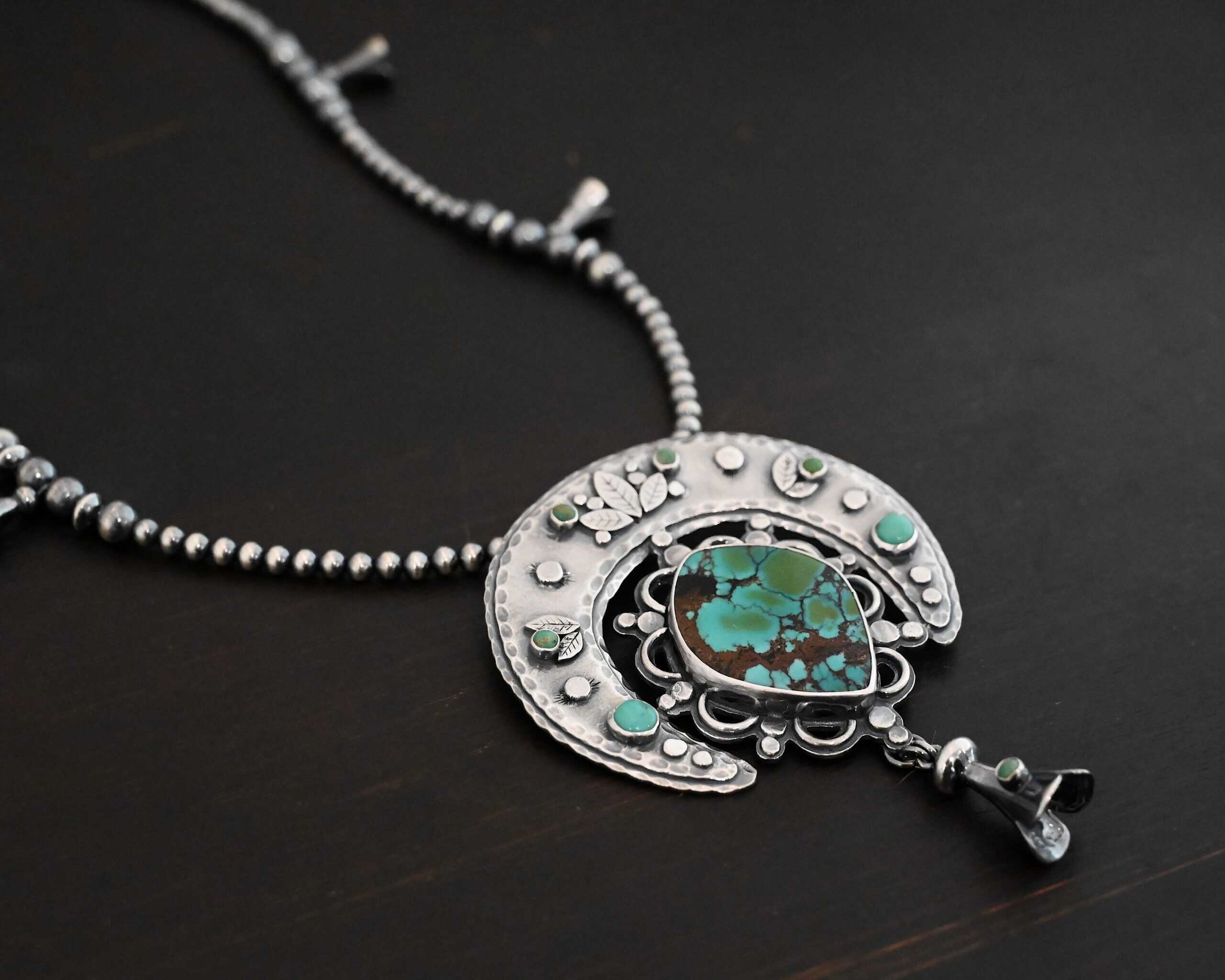 Wanderer Squash Blossom Necklace Photo of the "Wanderer Squash Blossom" Necklace. It has a large turquoise necklace on a black background. It is a squash blossom necklace. There is a large turquoise in the center with a flower petal design around it. There are seven other small turquoise on the horseshoe shaped silver piece going around the flower design of the turquoise. This piece has silver leaves and hammered silver dots on it as well. Hanging beneath the turquoise is a small cone of silver with cuts in it to make it look like an upside down flower petal. The chain is beaded and has the cone silver flower petals scattered around the chain.