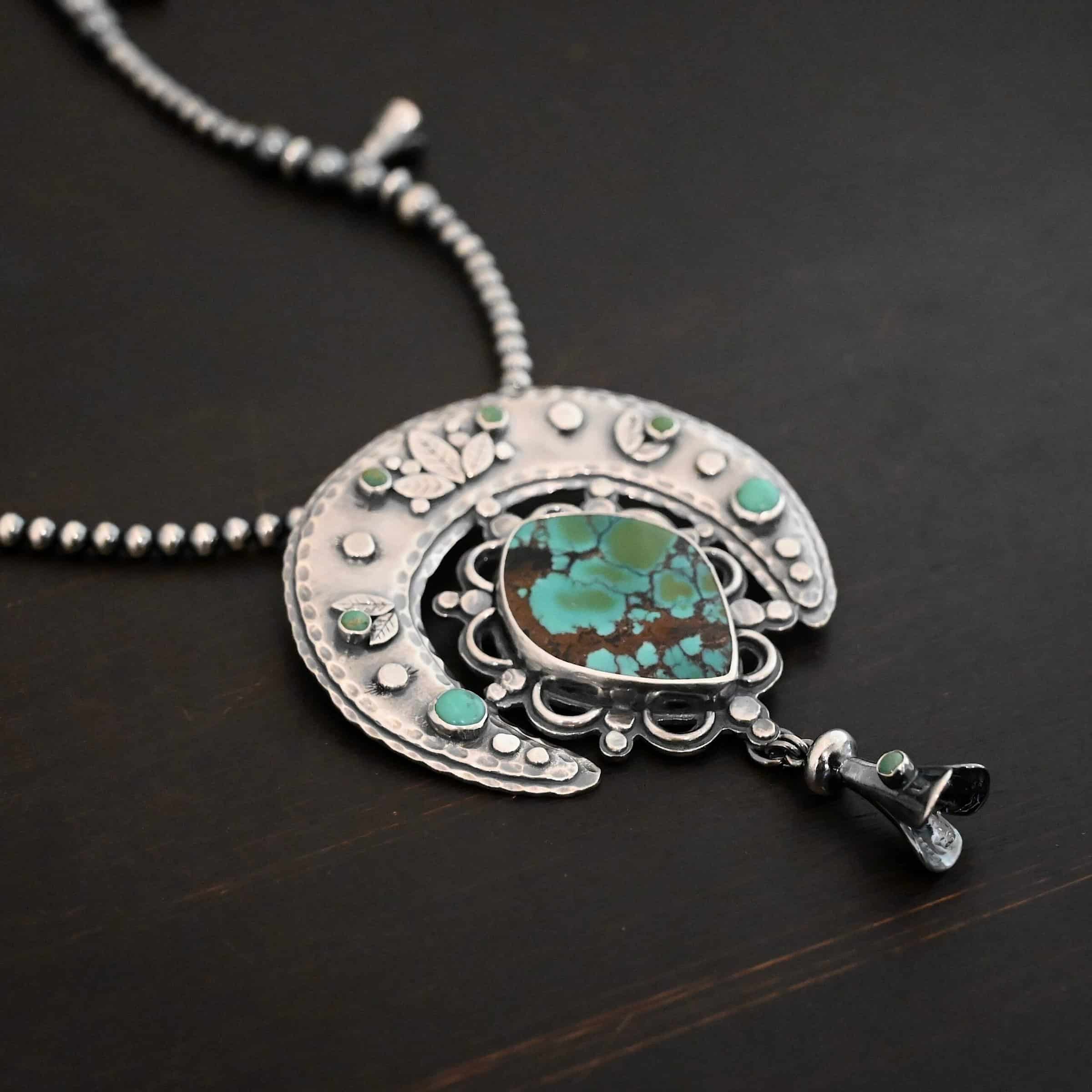 Wanderer Squash Blossom Necklace Photo of the "Wanderer Squash Blossom" Necklace. It has a large turquoise necklace on a black background. It is a squash blossom necklace. There is a large turquoise in the center with a flower petal design around it. There are seven other small turquoise on the horseshoe shaped silver piece going around the flower design of the turquoise. This piece has silver leaves and hammered silver dots on it as well. Hanging beneath the turquoise is a small cone of silver with cuts in it to make it look like an upside down flower petal. The chain is beaded and has the cone silver flower petals scattered around the chain.