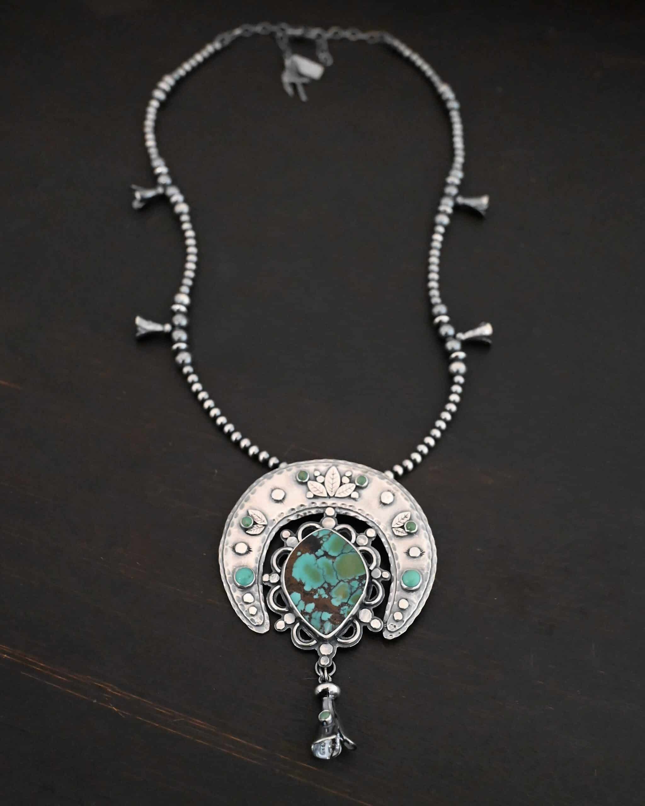 Wanderer Squash Blossom Necklace Photo of the "Wanderer Squash Blossom" Necklace. It has a large turquoise necklace on a black background. It is a squash blossom necklace. There is a large turquoise in the center with a flower petal design around it. There are seven other small turquoise on the horseshoe shaped silver piece going around the flower design of the turquoise. This piece has silver leaves and hammered silver dots on it as well. Hanging beneath the turquoise is a small cone of silver with cuts in it to make it look like an upside down flower petal. The chain is beaded and has the cone silver flower petals scattered around the chain.
