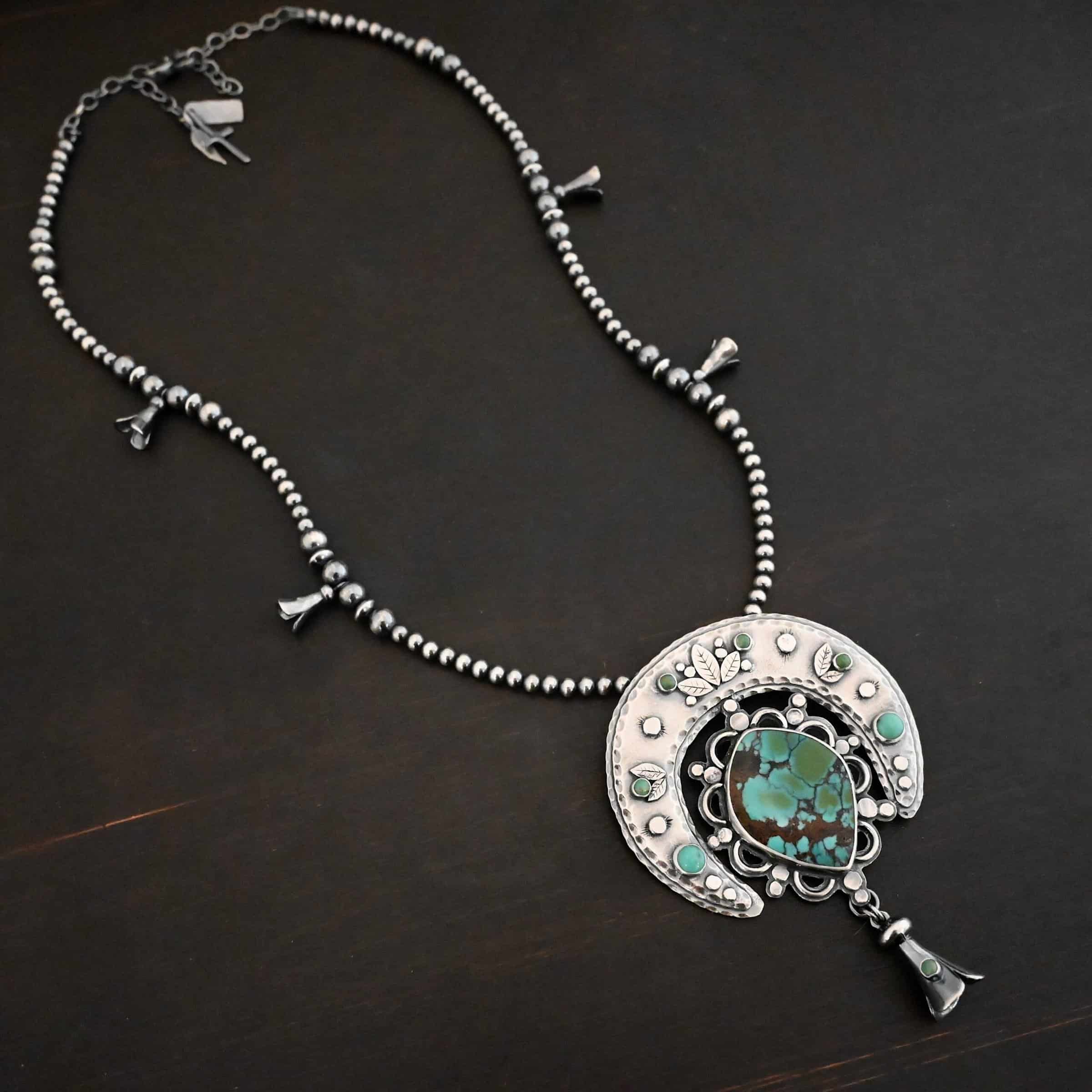 Wanderer Squash Blossom Necklace Photo of the "Wanderer Squash Blossom" Necklace. It has a large turquoise necklace on a black background. It is a squash blossom necklace. There is a large turquoise in the center with a flower petal design around it. There are seven other small turquoise on the horseshoe shaped silver piece going around the flower design of the turquoise. This piece has silver leaves and hammered silver dots on it as well. Hanging beneath the turquoise is a small cone of silver with cuts in it to make it look like an upside down flower petal. The chain is beaded and has the cone silver flower petals scattered around the chain.