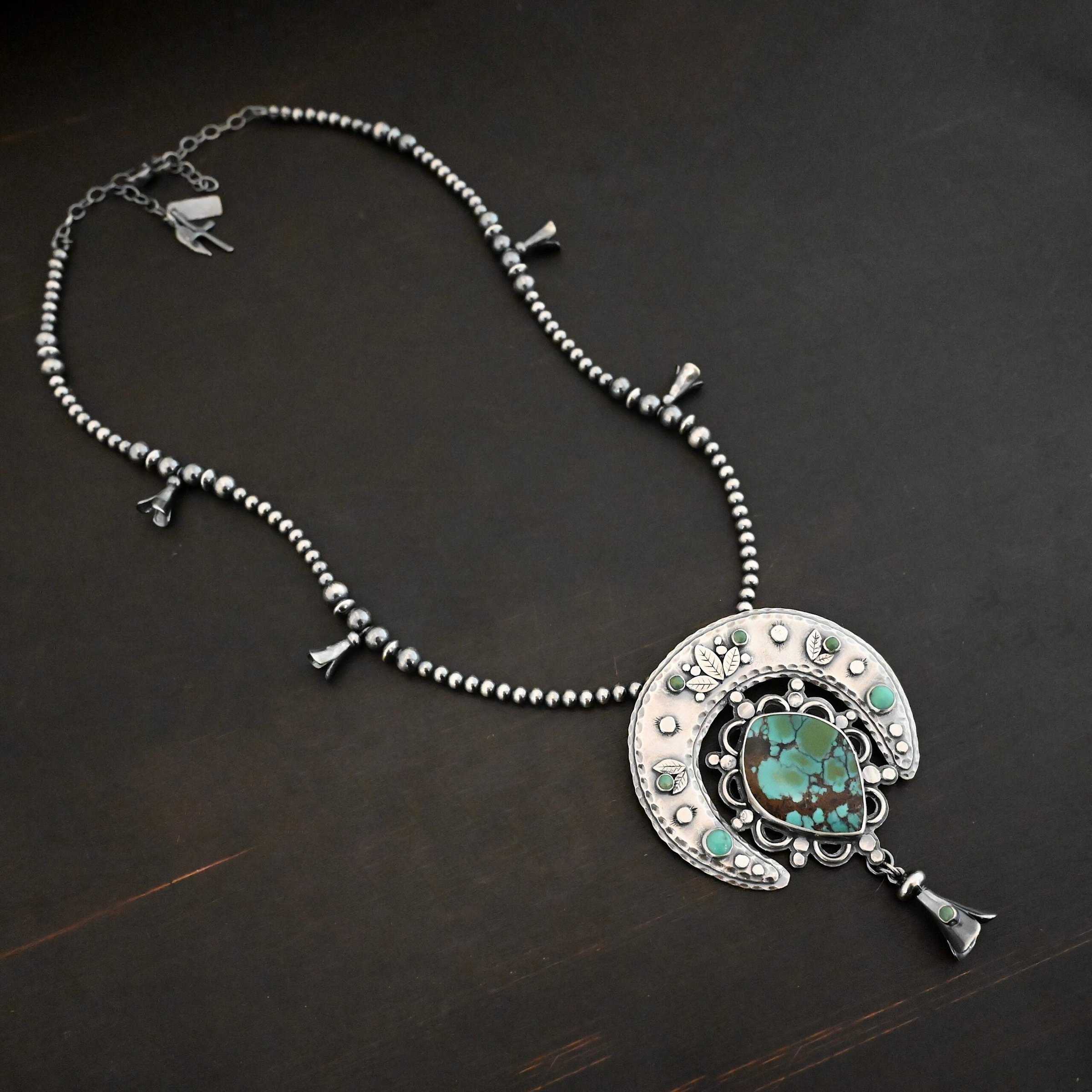Wanderer Squash Blossom Necklace Photo of the "Wanderer Squash Blossom" Necklace. It has a large turquoise necklace on a black background. It is a squash blossom necklace. There is a large turquoise in the center with a flower petal design around it. There are seven other small turquoise on the horseshoe shaped silver piece going around the flower design of the turquoise. This piece has silver leaves and hammered silver dots on it as well. Hanging beneath the turquoise is a small cone of silver with cuts in it to make it look like an upside down flower petal. The chain is beaded and has the cone silver flower petals scattered around the chain.