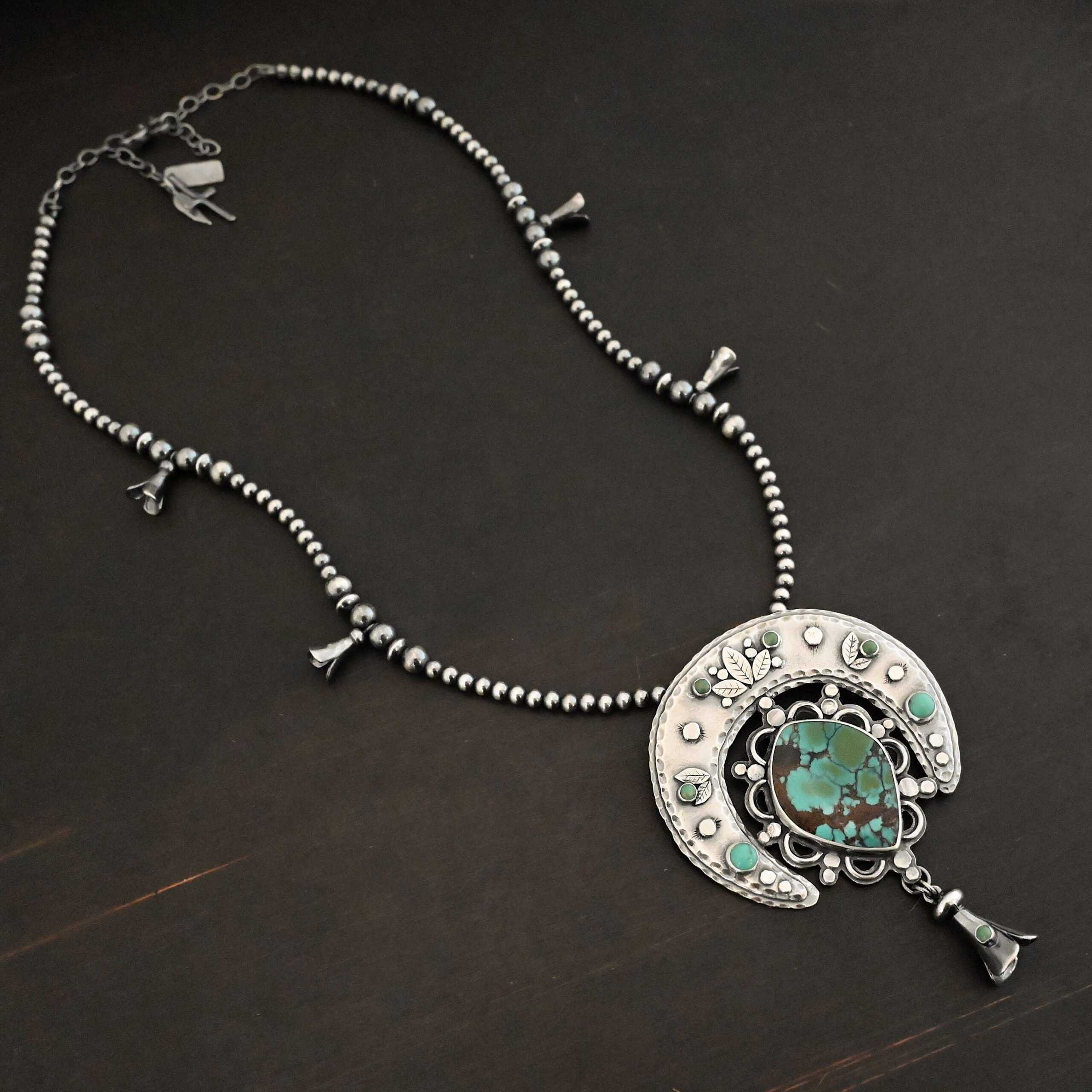 Wanderer Squash Blossom Necklace Photo of the "Wanderer Squash Blossom" Necklace. It has a large turquoise necklace on a black background. It is a squash blossom necklace. There is a large turquoise in the center with a flower petal design around it. There are seven other small turquoise on the horseshoe shaped silver piece going around the flower design of the turquoise. This piece has silver leaves and hammered silver dots on it as well. Hanging beneath the turquoise is a small cone of silver with cuts in it to make it look like an upside down flower petal. The chain is beaded and has the cone silver flower petals scattered around the chain.
