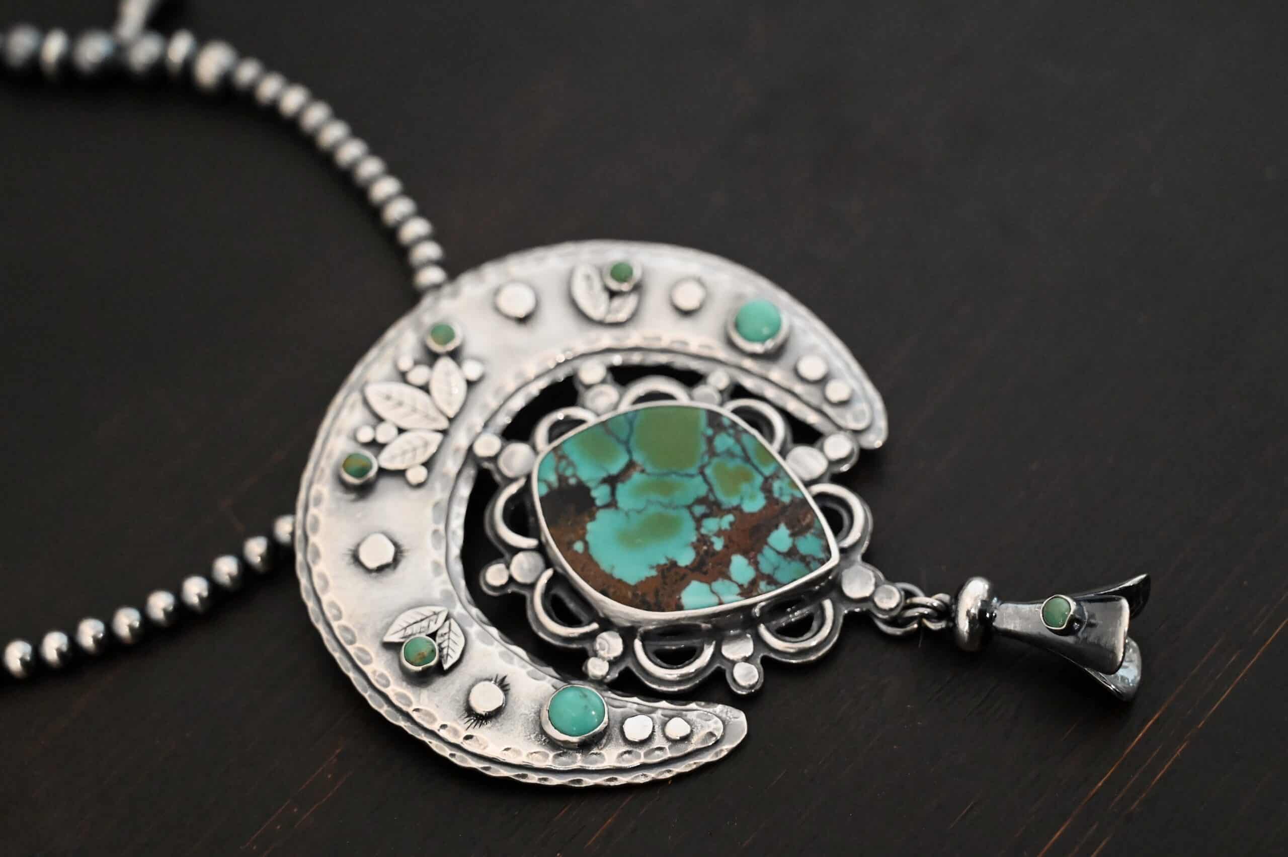 Wanderer Squash Blossom Necklace Photo of the "Wanderer Squash Blossom" Necklace. It has a large turquoise necklace on a black background. It is a squash blossom necklace. There is a large turquoise in the center with a flower petal design around it. There are seven other small turquoise on the horseshoe shaped silver piece going around the flower design of the turquoise. This piece has silver leaves and hammered silver dots on it as well. Hanging beneath the turquoise is a small cone of silver with cuts in it to make it look like an upside down flower petal. Necklace is laying against a dark background.