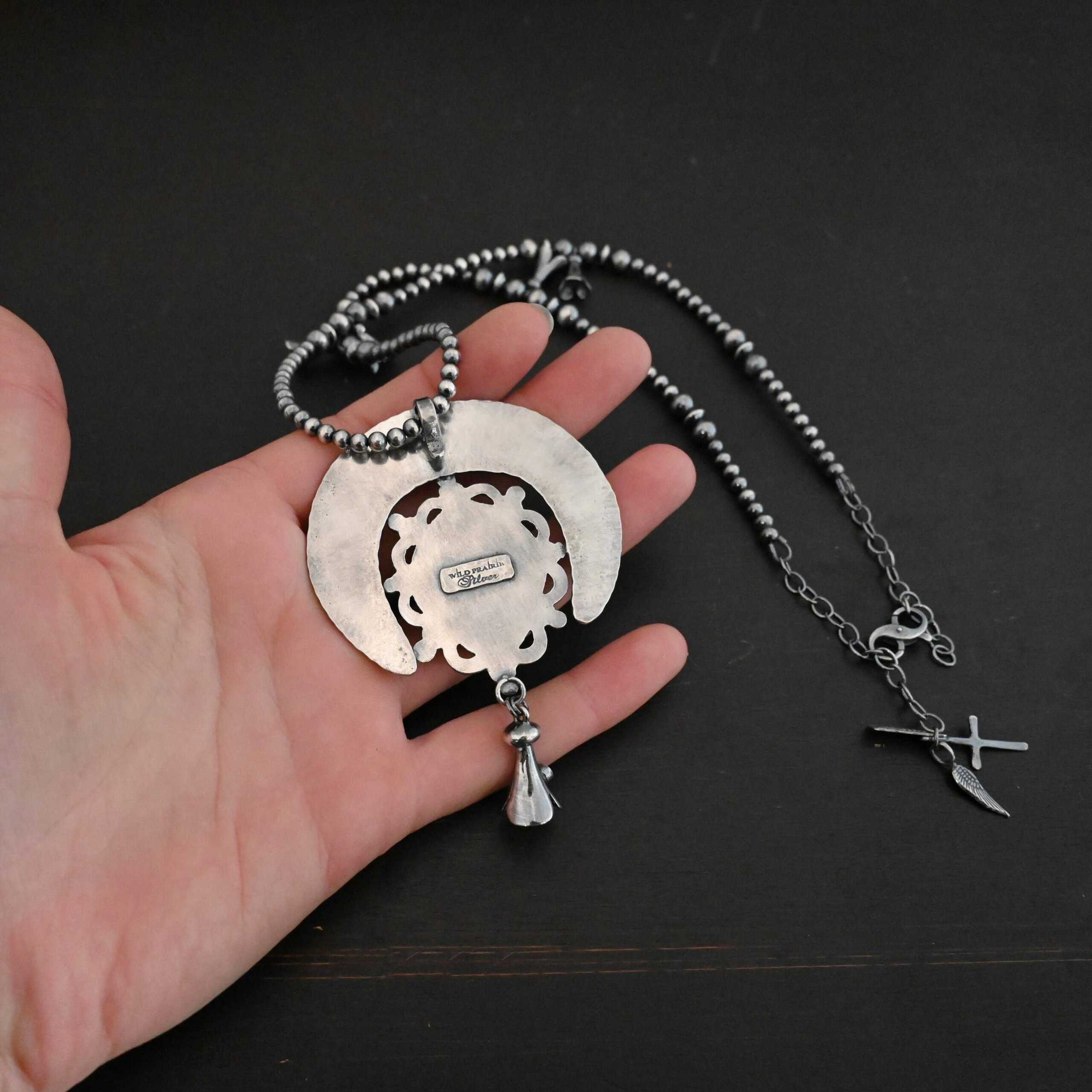 Wanderer Squash Blossom Necklace Photo of the back of the "Wanderer Squash Blossom" Necklace. Made out of sterling silver. The back of the necklace starts with a horseshoe shape piece of silver, and a large ornately carved piece of silver around the edges attached to the front of it. There is a cone shaped flower attached to the bottom of the pendant with a soldered loop. There is a small rectangle on the back that has the words "Wild Prairie Silver" stamped into it. The chain of the necklace is beaded and also has cone shaped flowers scattered around the chain. There is a cross and angel wing charm on the necklace extension. Necklace is laying in the palm of a hand to demonstrate size.