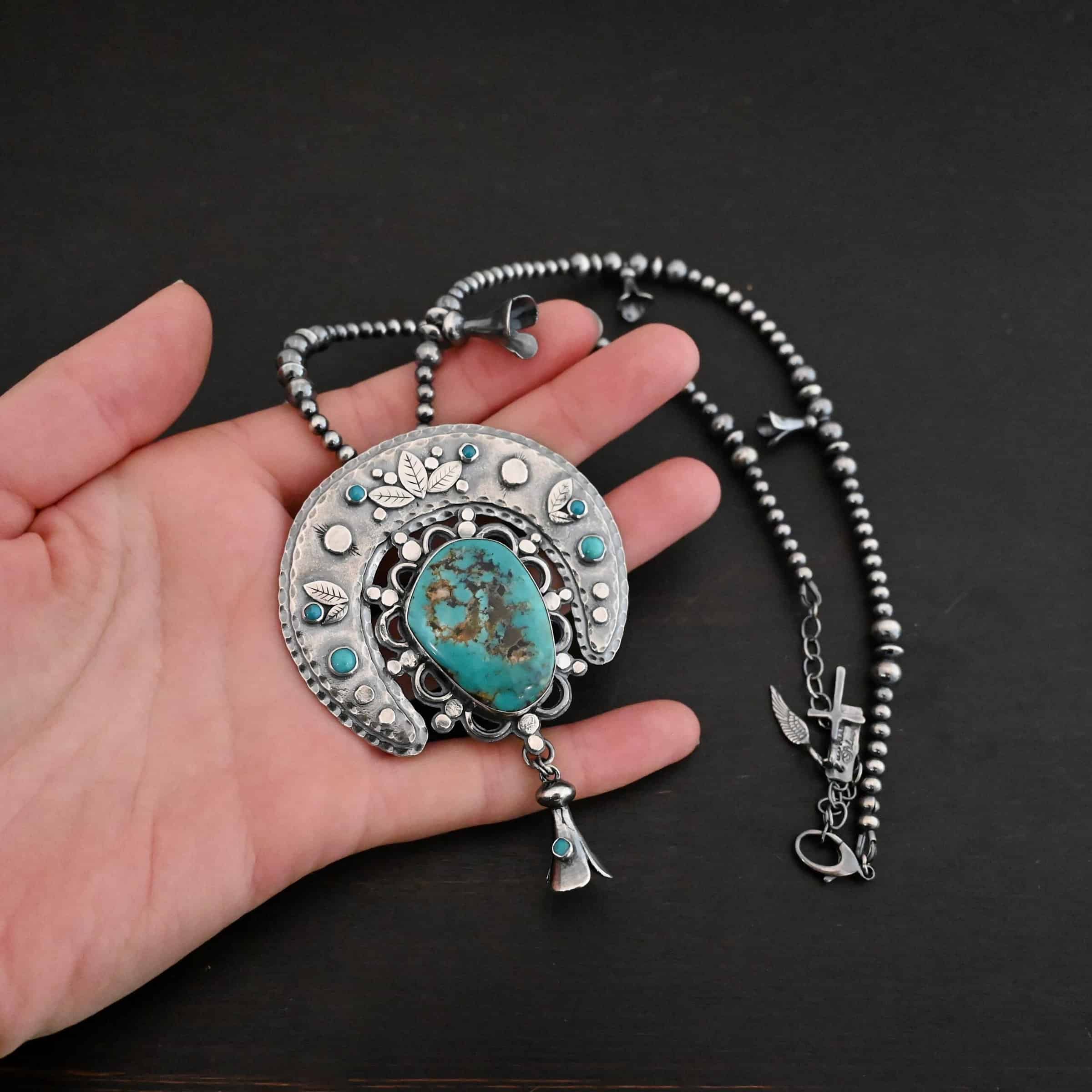 Photo of the "Dreamer Squash Blossom" Necklace. It has a large turquoise necklace on a black background. It is a squash blossom necklace. There is a large turquoise in the center with a flower petal design around it. There are seven other small turquoise on the horseshoe shaped silver piece going around the flower design of the turquoise. This piece has silver leaves and hammered silver dots on it as well. Hanging beneath the turquoise is a small cone of silver with cuts in it to make it look like an upside down flower petal. The chain is beaded and has the cone silver flower petals scattered around the chain. Necklace is lying against the palm of a hand to demonstrate size.