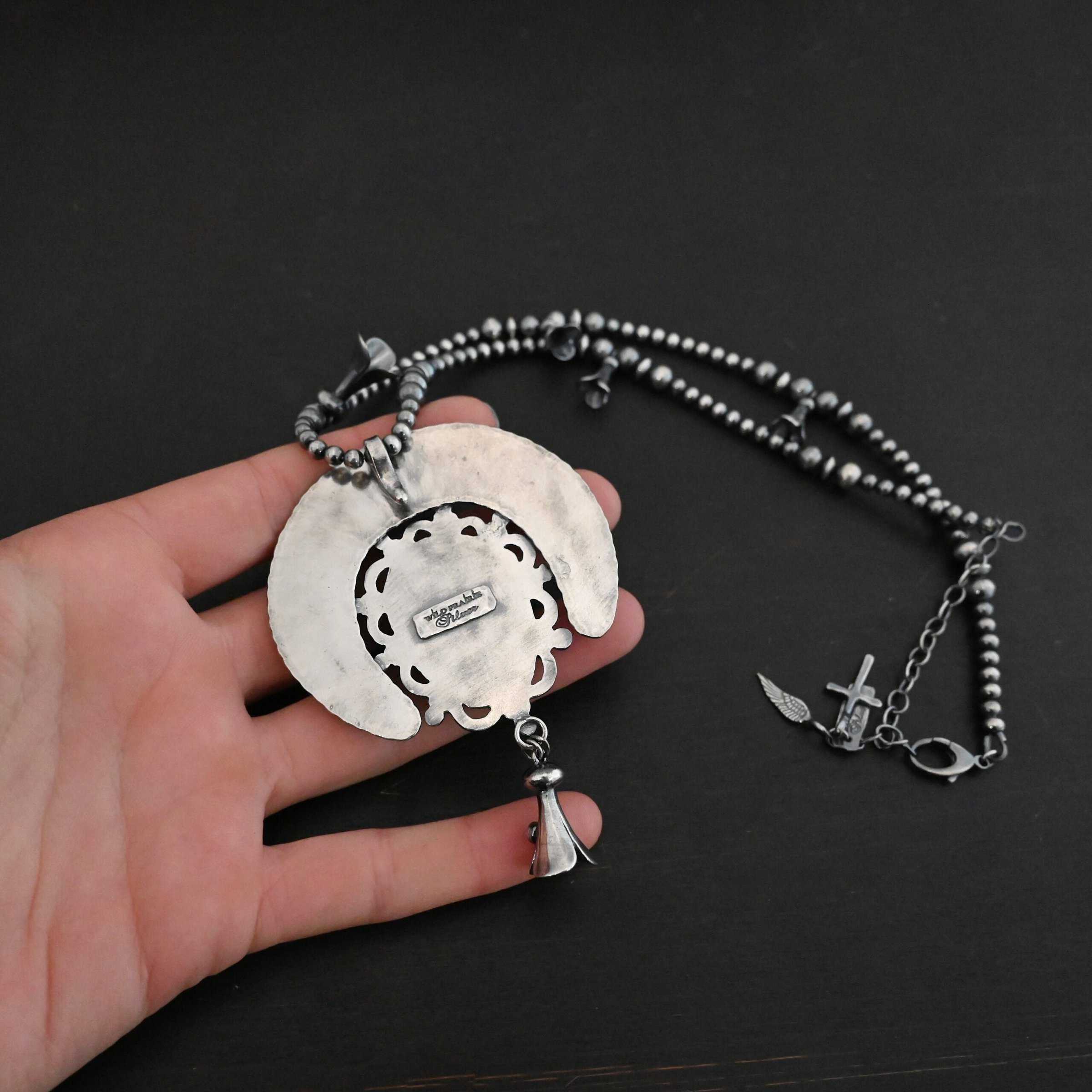 Photo of the back of the "Dreamer Squash Blossom" Necklace. Made out of sterling silver. The back of the necklace starts with a horseshoe shape piece of silver, and a large ornately carved piece of silver around the edges attached to the front of it. There is a cone shaped flower attached to the bottom of the pendant with a soldered loop. There is a small rectangle on the back that has the words "Wild Prairie Silver" stamped into it. The chain of the necklace is beaded and also has cone shaped flowers scattered around the chain. There is a cross and angel wing charm on the necklace extension. Necklace is laying in the palm of a hand to demonstrate size.
