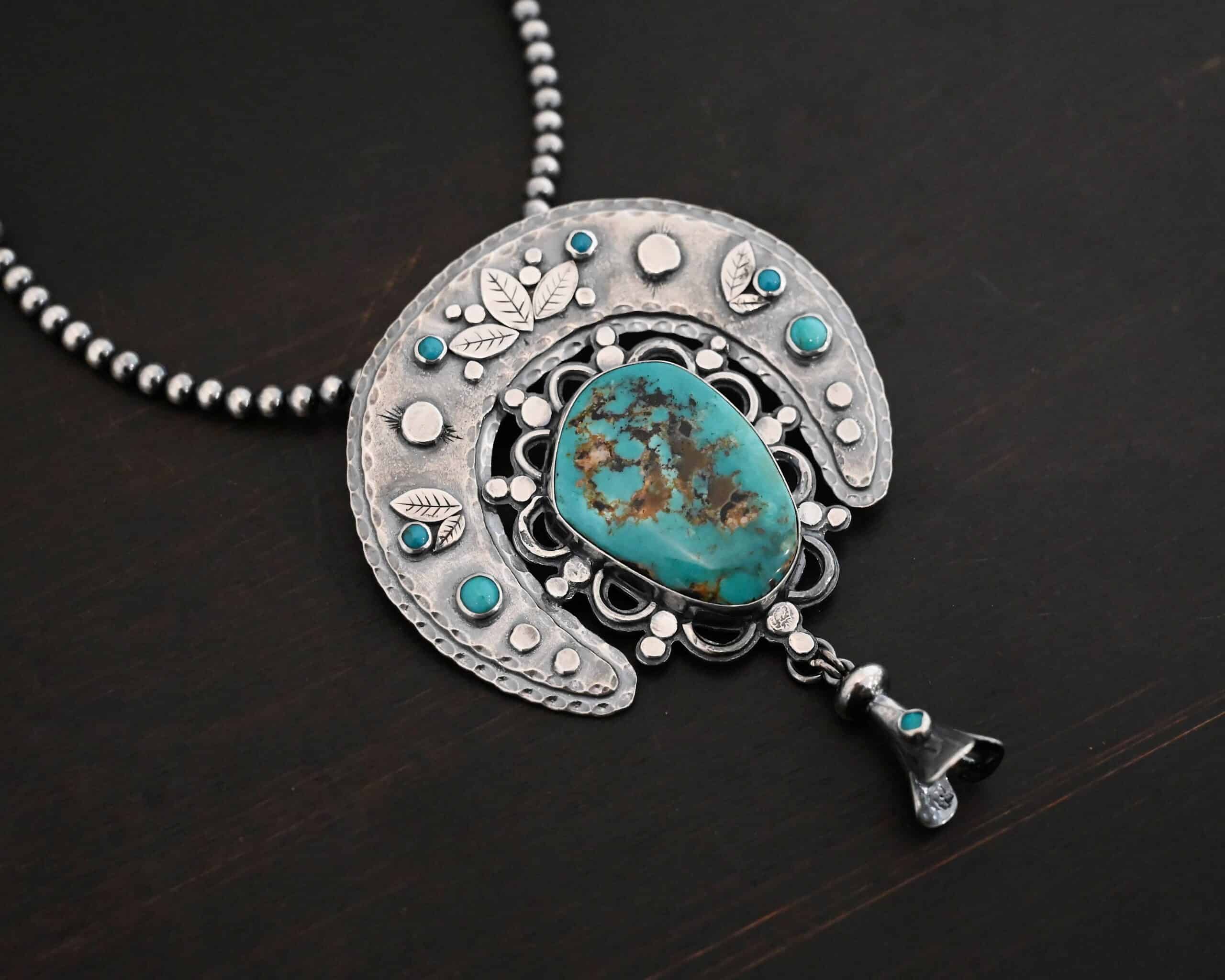 Photo of the "Dreamer Squash Blossom" Necklace. It has a large turquoise necklace on a black background. It is a squash blossom necklace. There is a large turquoise in the center with a flower petal design around it. There are seven other small turquoise on the horseshoe shaped silver piece going around the flower design of the turquoise. This piece has silver leaves and hammered silver dots on it as well. Hanging beneath the turquoise is a small cone of silver with cuts in it to make it look like an upside down flower petal. The chain is beaded and has the cone silver flower petals scattered around the chain.