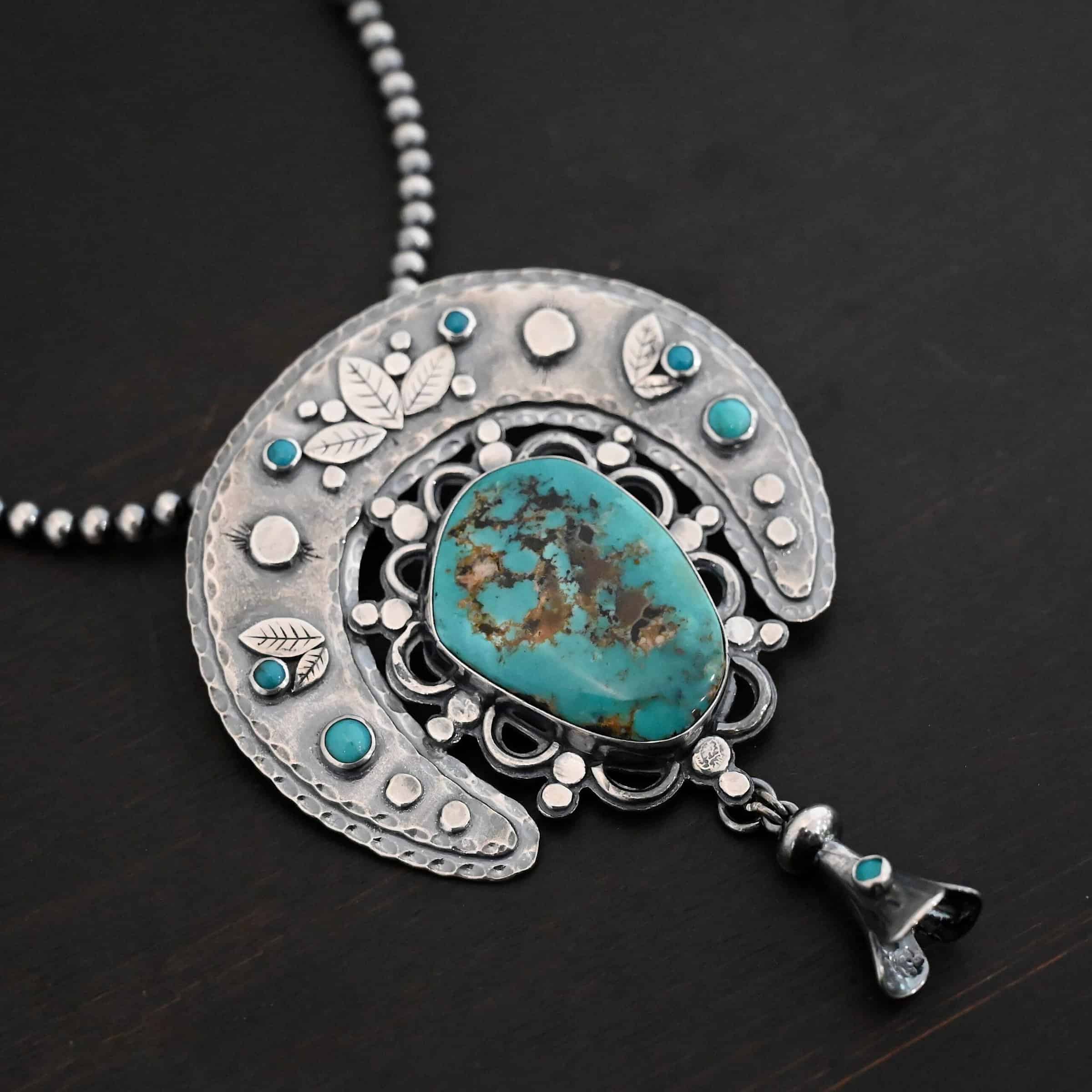 Photo of the "Dreamer Squash Blossom" Necklace. It has a large turquoise necklace on a black background. It is a squash blossom necklace. There is a large turquoise in the center with a flower petal design around it. There are seven other small turquoise on the horseshoe shaped silver piece going around the flower design of the turquoise. This piece has silver leaves and hammered silver dots on it as well. Hanging beneath the turquoise is a small cone of silver with cuts in it to make it look like an upside down flower petal. The chain is beaded and has the cone silver flower petals scattered around the chain.