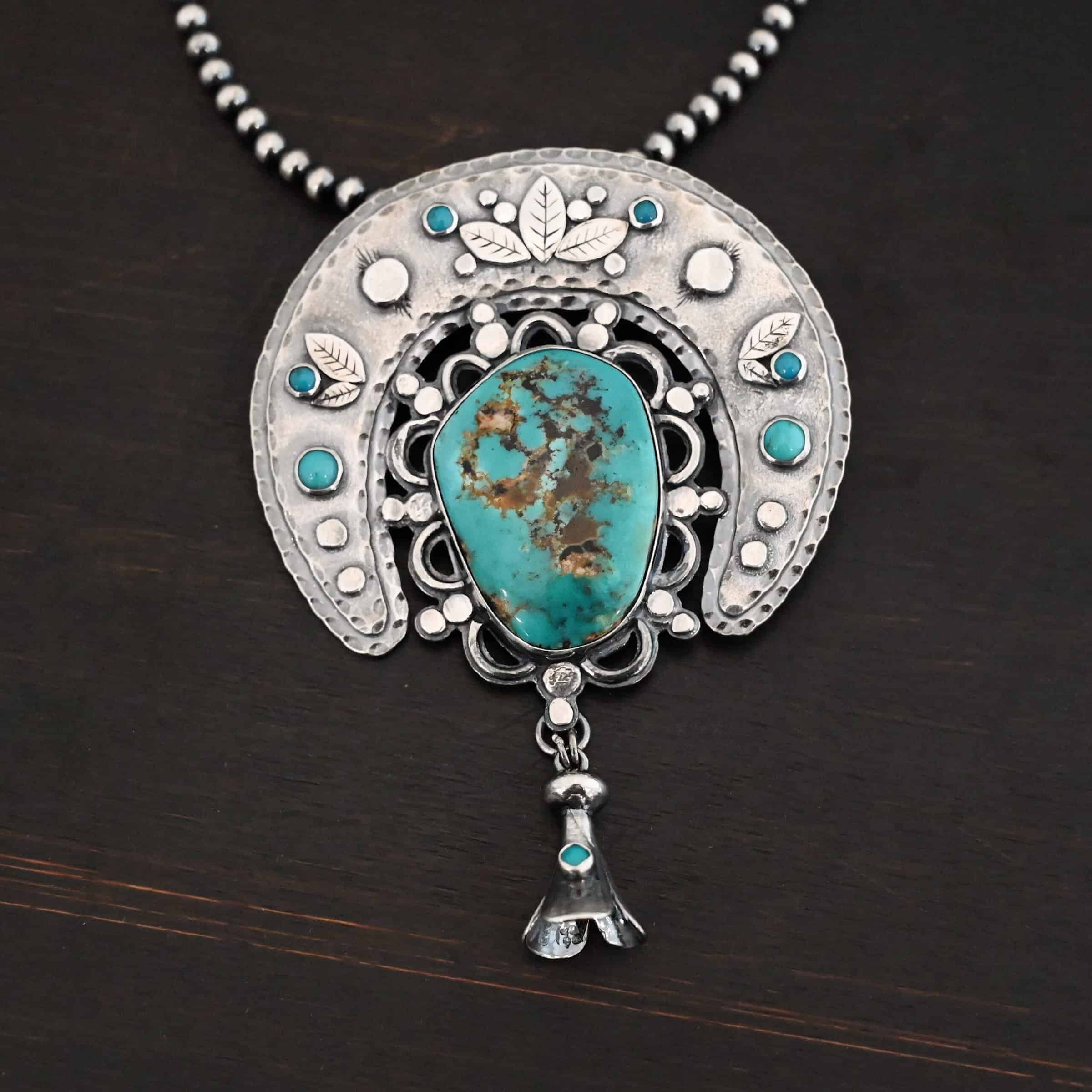 Photo of the "Dreamer Squash Blossom" Necklace. It has a large turquoise necklace on a black background. It is a squash blossom necklace. There is a large turquoise in the center with a flower petal design around it. There are seven other small turquoise on the horseshoe shaped silver piece going around the flower design of the turquoise. This piece has silver leaves and hammered silver dots on it as well. Hanging beneath the turquoise is a small cone of silver with cuts in it to make it look like an upside down flower petal. The chain is beaded and has the cone silver flower petals scattered around the chain.