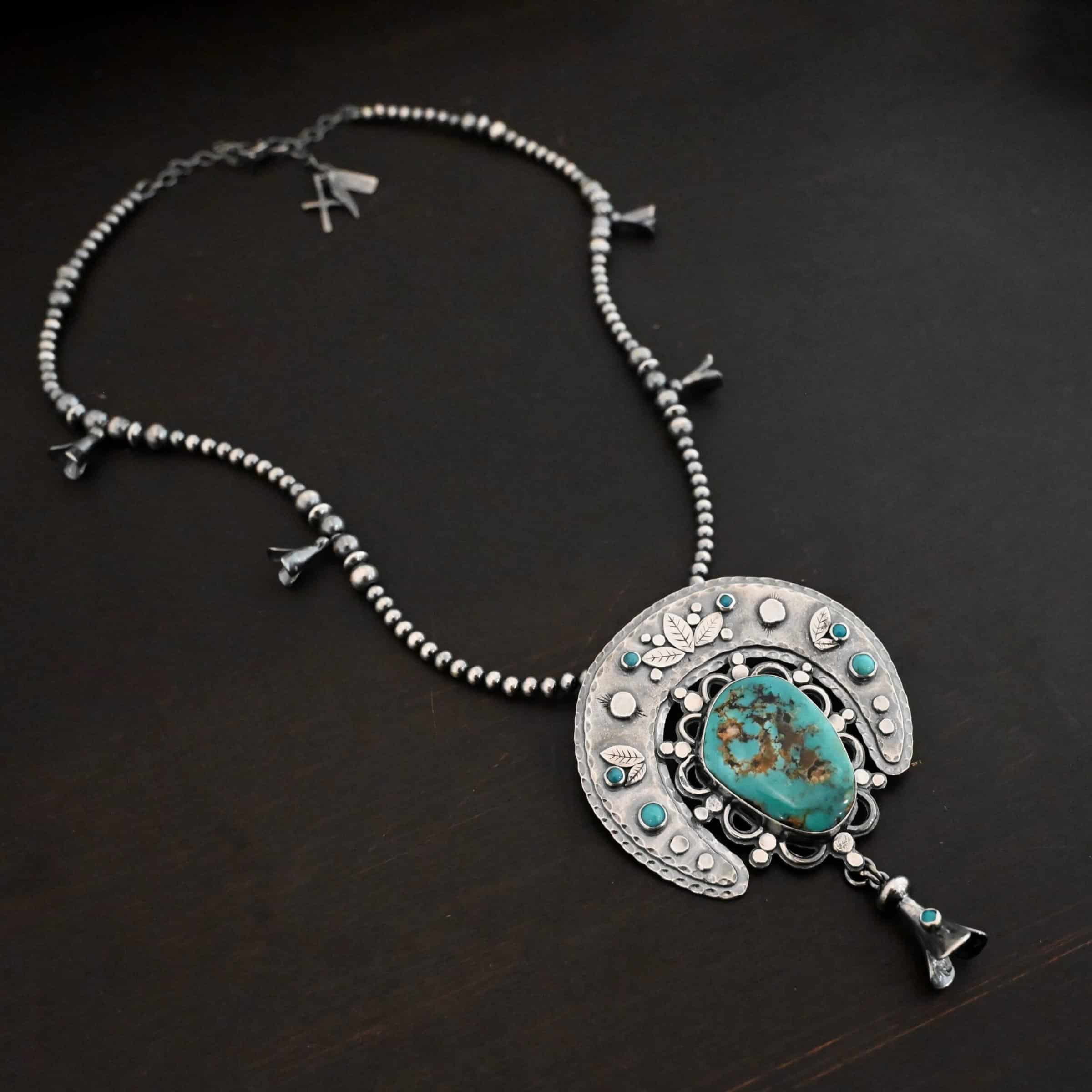 Photo of the "Dreamer Squash Blossom" Necklace. It has a large turquoise necklace on a black background. It is a squash blossom necklace. There is a large turquoise in the center with a flower petal design around it. There are seven other small turquoise on the horseshoe shaped silver piece going around the flower design of the turquoise. This piece has silver leaves and hammered silver dots on it as well. Hanging beneath the turquoise is a small cone of silver with cuts in it to make it look like an upside down flower petal. The chain is beaded and has the cone silver flower petals scattered around the chain.