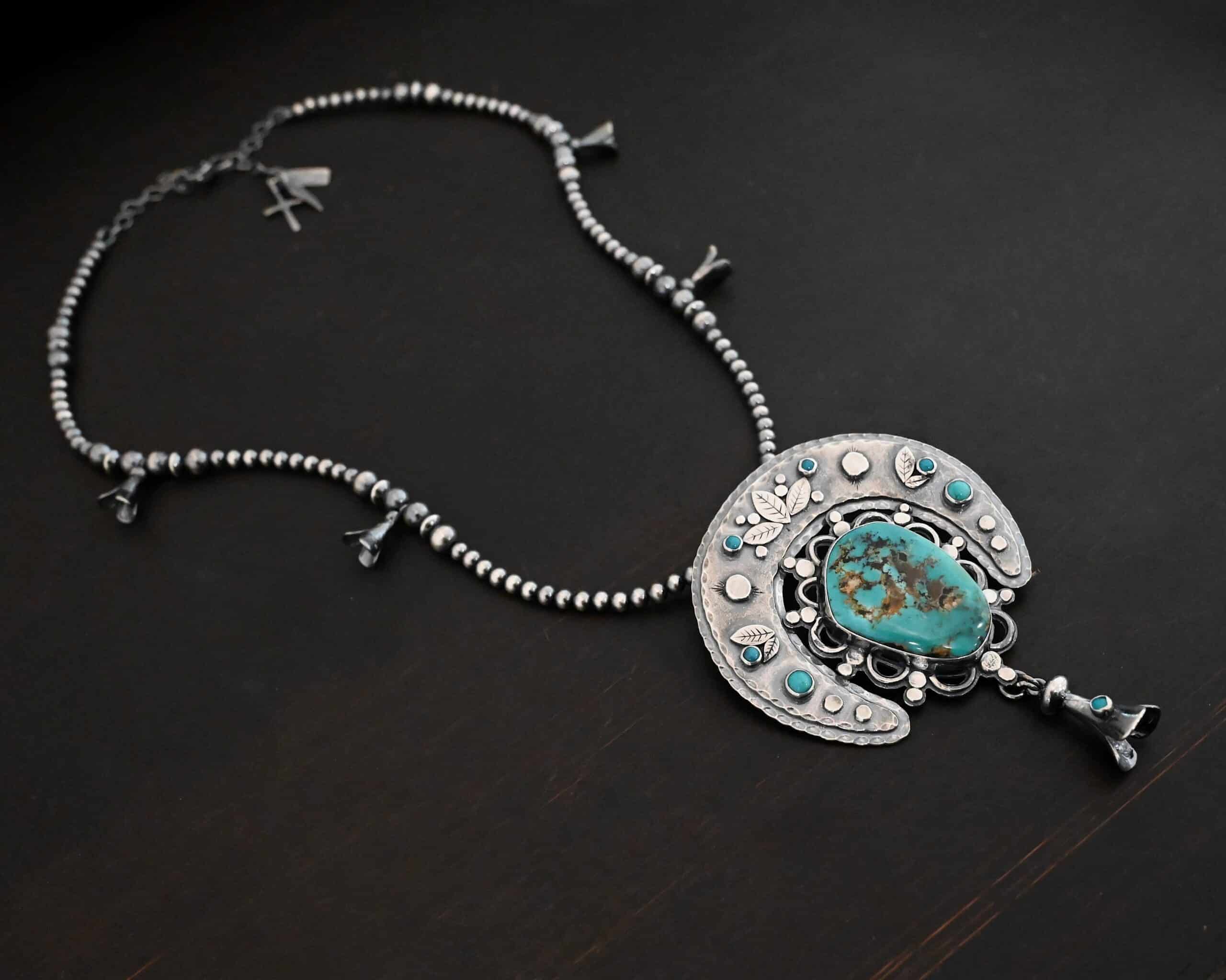 Photo of the "Dreamer Squash Blossom" Necklace. It has a large turquoise necklace on a black background. It is a squash blossom necklace. There is a large turquoise in the center with a flower petal design around it. There are seven other small turquoise on the horseshoe shaped silver piece going around the flower design of the turquoise. This piece has silver leaves and hammered silver dots on it as well. Hanging beneath the turquoise is a small cone of silver with cuts in it to make it look like an upside down flower petal. The chain is beaded and has the cone silver flower petals scattered around the chain.