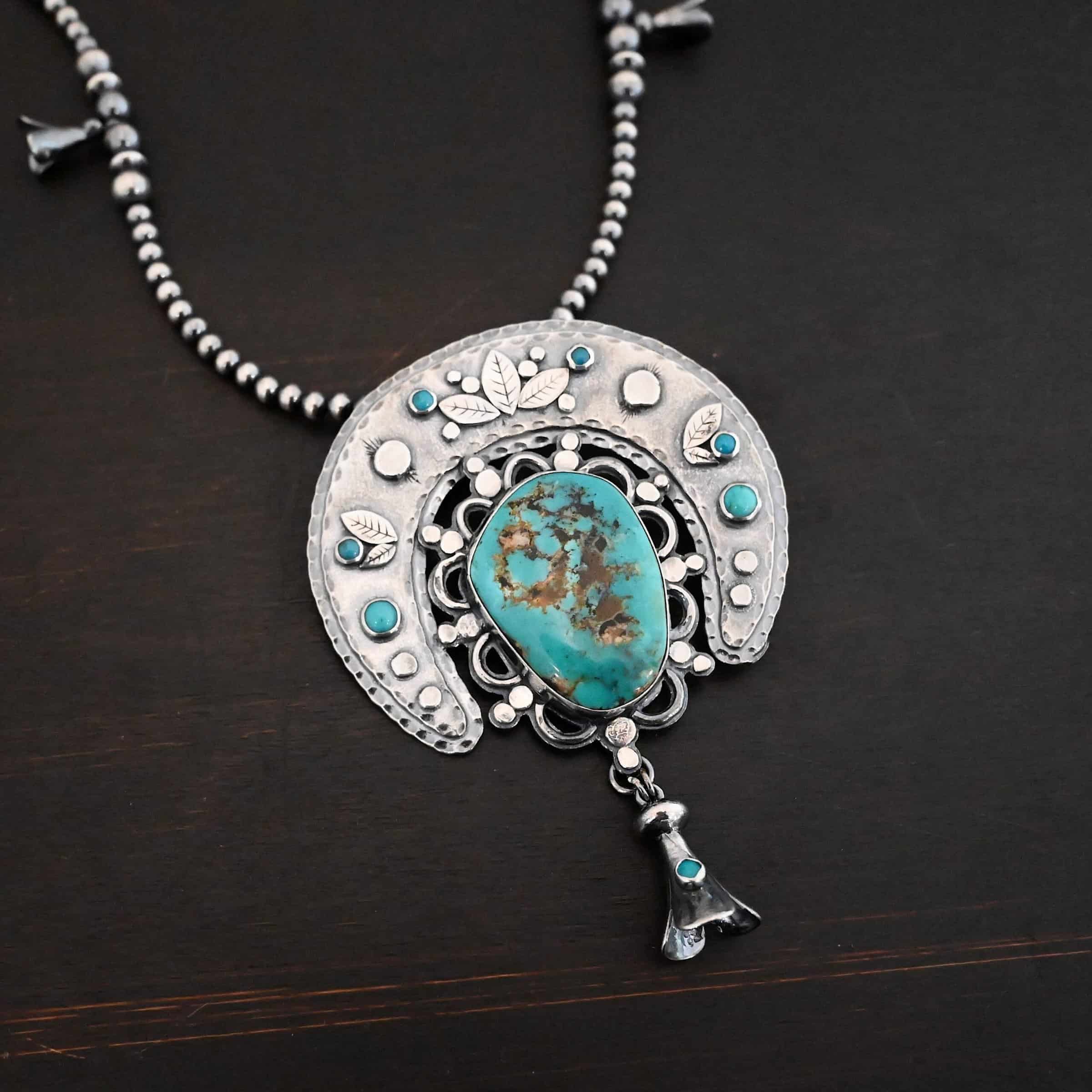 Photo of the "Dreamer Squash Blossom" Necklace. It has a large turquoise necklace on a black background. It is a squash blossom necklace. There is a large turquoise in the center with a flower petal design around it. There are seven other small turquoise on the horseshoe shaped silver piece going around the flower design of the turquoise. This piece has silver leaves and hammered silver dots on it as well. Hanging beneath the turquoise is a small cone of silver with cuts in it to make it look like an upside down flower petal. The chain is beaded and has the cone silver flower petals scattered around the chain.