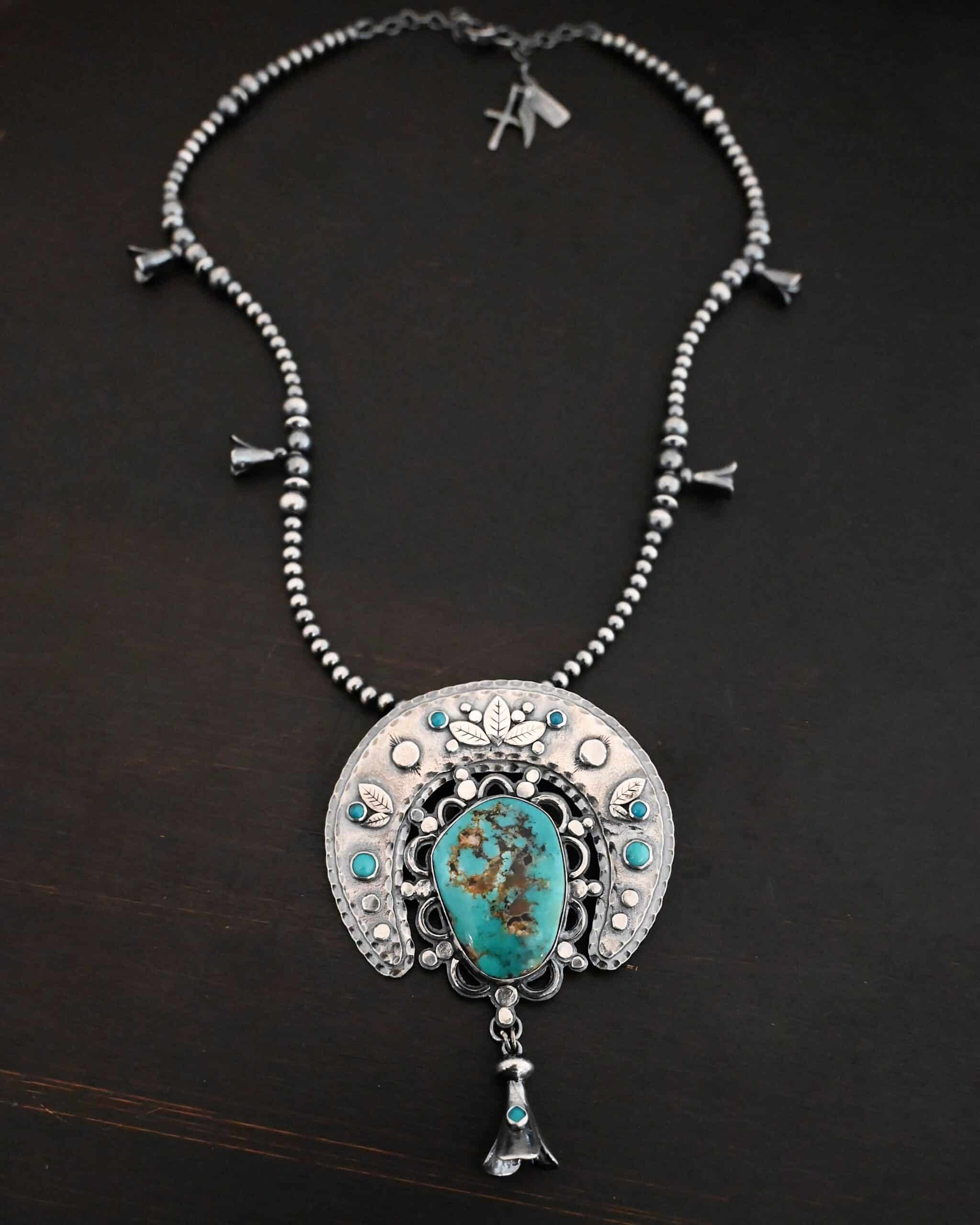 Photo of the "Dreamer Squash Blossom" Necklace. It has a large turquoise necklace on a black background. It is a squash blossom necklace. There is a large turquoise in the center with a flower petal design around it. There are seven other small turquoise on the horseshoe shaped silver piece going around the flower design of the turquoise. This piece has silver leaves and hammered silver dots on it as well. Hanging beneath the turquoise is a small cone of silver with cuts in it to make it look like an upside down flower petal. The chain is beaded and has the cone silver flower petals scattered around the chain.
