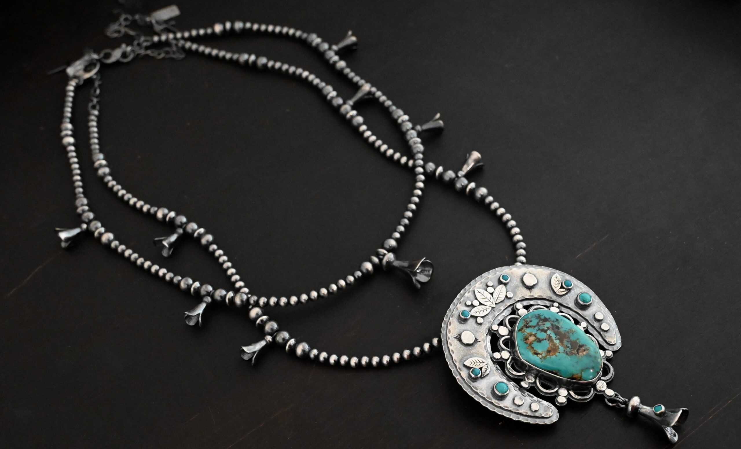 Photo of the "Dreamer Squash Blossom" Necklace. It has a large turquoise necklace on a black background. It is a squash blossom necklace. There is a large turquoise in the center with a flower petal design around it. There are seven other small turquoise on the horseshoe shaped silver piece going around the flower design of the turquoise. This piece has silver leaves and hammered silver dots on it as well. Hanging beneath the turquoise is a small cone of silver with cuts in it to make it look like an upside down flower petal. The chain is beaded and has the cone silver flower petals scattered around the chain. There is another beaded chain with cone flower petals on top of the necklace that goes with the necklace.