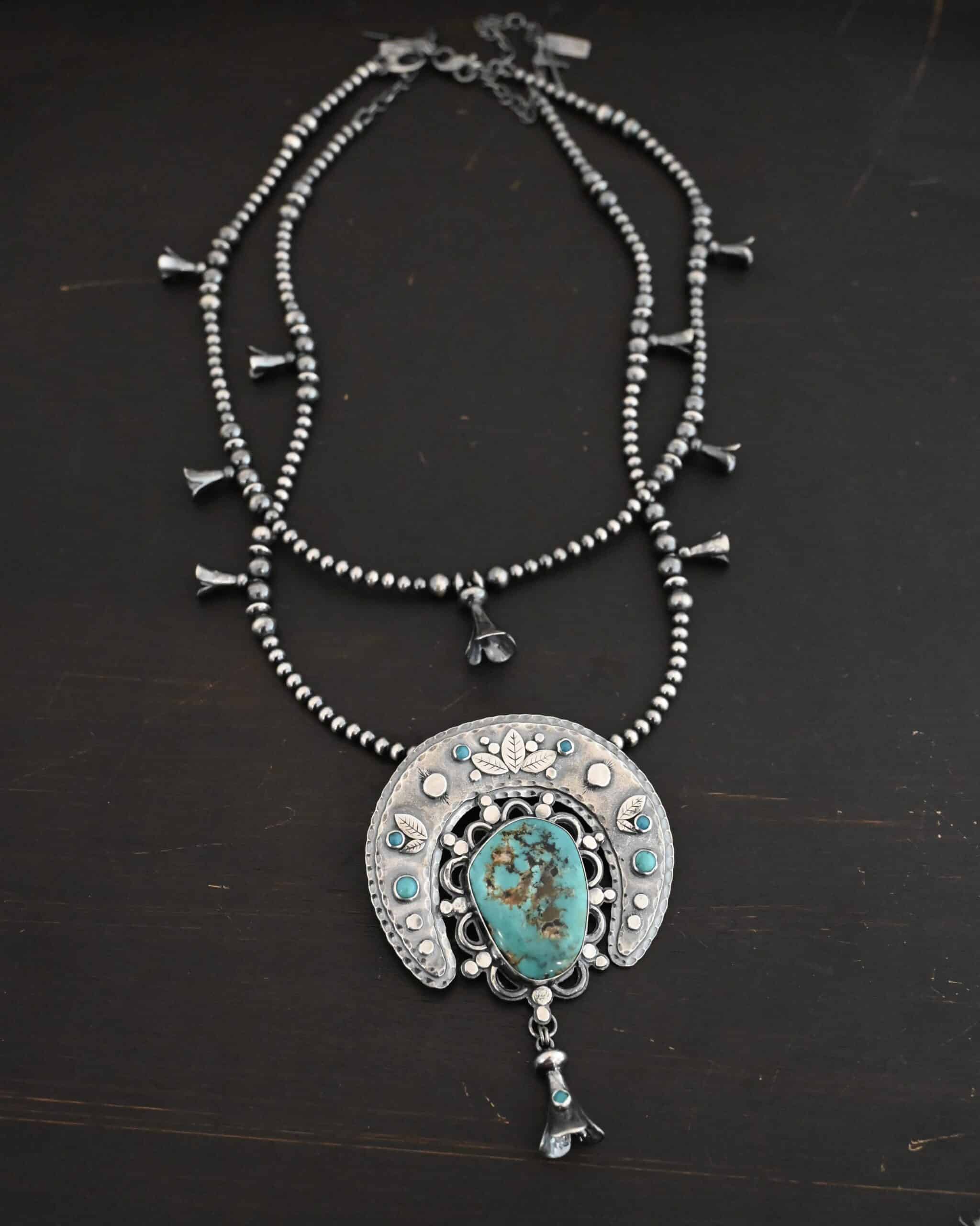 Photo of the "Wanderer Squash Blossom" Necklace. It has a large turquoise necklace on a black background. It is a squash blossom necklace. There is a large turquoise in the center with a flower petal design around it. There are seven other small turquoise on the horseshoe shaped silver piece going around the flower design of the turquoise. This piece has silver leaves and hammered silver dots on it as well. Hanging beneath the turquoise is a small cone of silver with cuts in it to make it look like an upside down flower petal. The chain is beaded and has the cone silver flower petals scattered around the chain. There is another beaded chain with cone flower petals on top of the necklace that goes with the necklace.