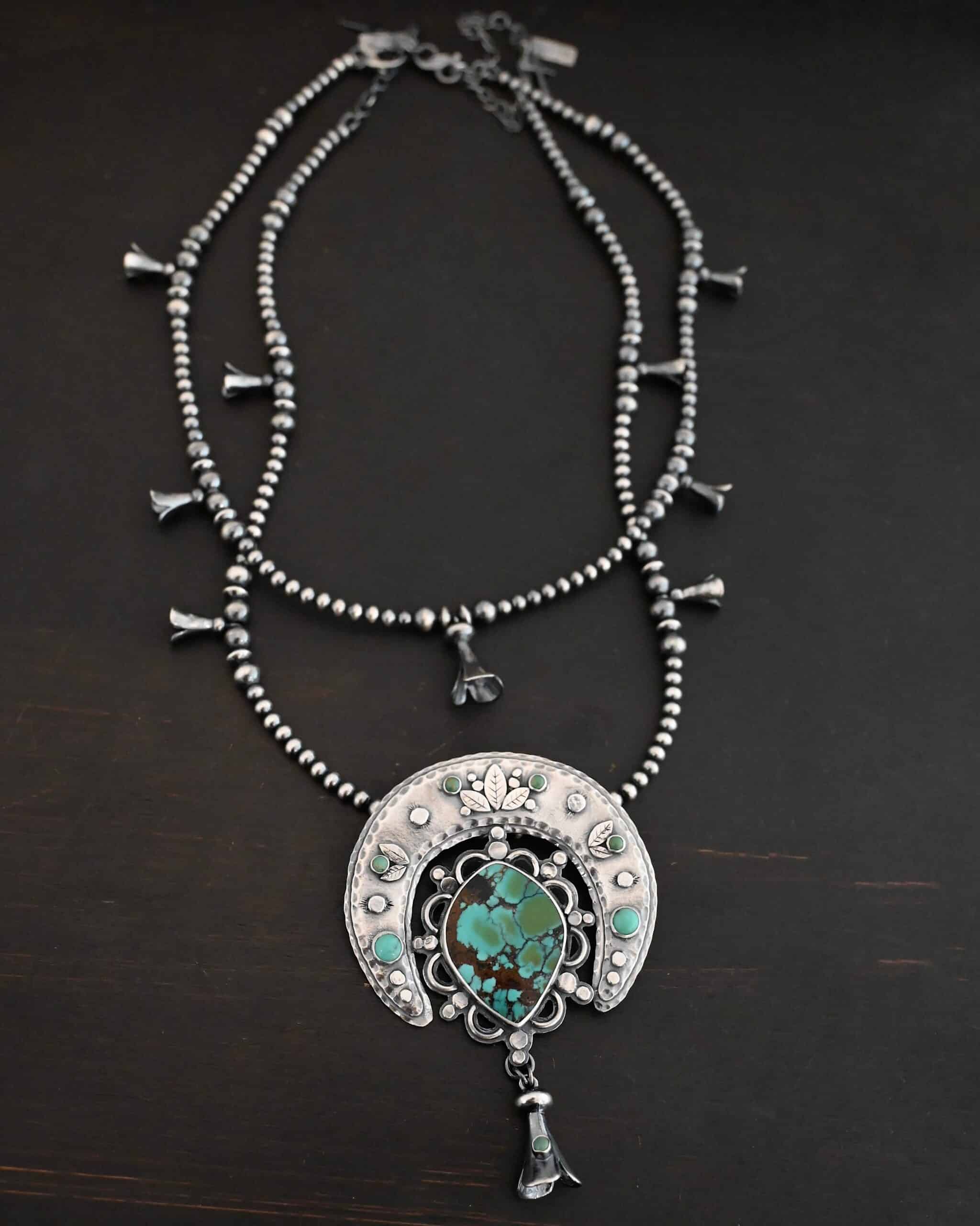 Wanderer Squash Blossom Necklace Photo of the "Wanderer Squash Blossom" Necklace. It has a large turquoise necklace on a black background. It is a squash blossom necklace. There is a large turquoise in the center with a flower petal design around it. There are seven other small turquoise on the horseshoe shaped silver piece going around the flower design of the turquoise. This piece has silver leaves and hammered silver dots on it as well. Hanging beneath the turquoise is a small cone of silver with cuts in it to make it look like an upside down flower petal. The chain is beaded and has the cone silver flower petals scattered around the chain. There is another beaded chain with cone flower petals on top of the necklace that goes with the necklace.