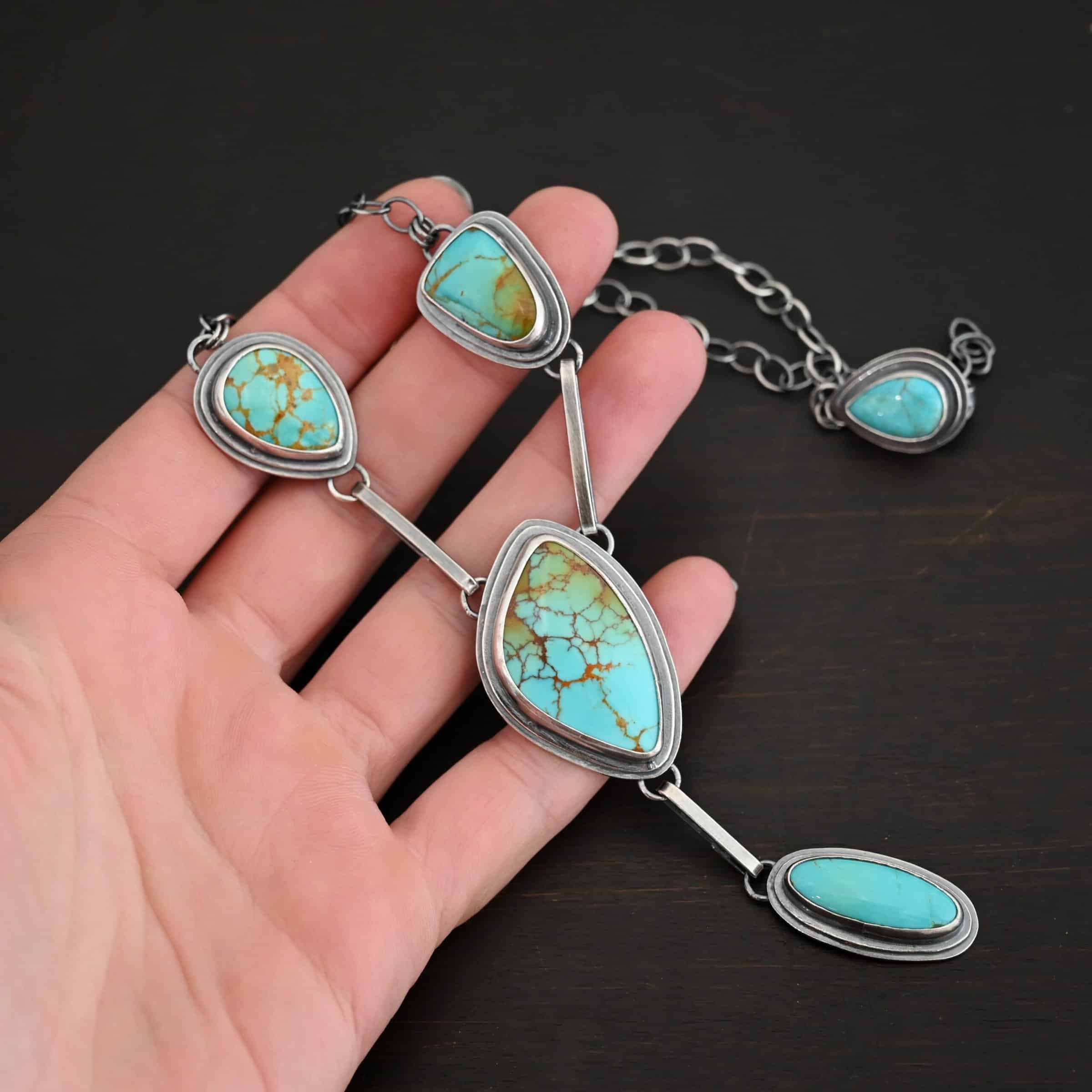 Gone with the Wind Necklace Photo of "Gone With The Wind" Necklace. This is a large turquoise necklace. The necklace has five turquoise, one on the left side, one on the right side, and two in the center. The two in the center are one on top of the other. There is a turquoise on the clasp of the necklace as well. Each turquoise is connected by a small silver bar and each bar is connected to loops on the backs of the settings for the turquoise. Necklace is laying against the fingertips of a hand to demonstrate size.