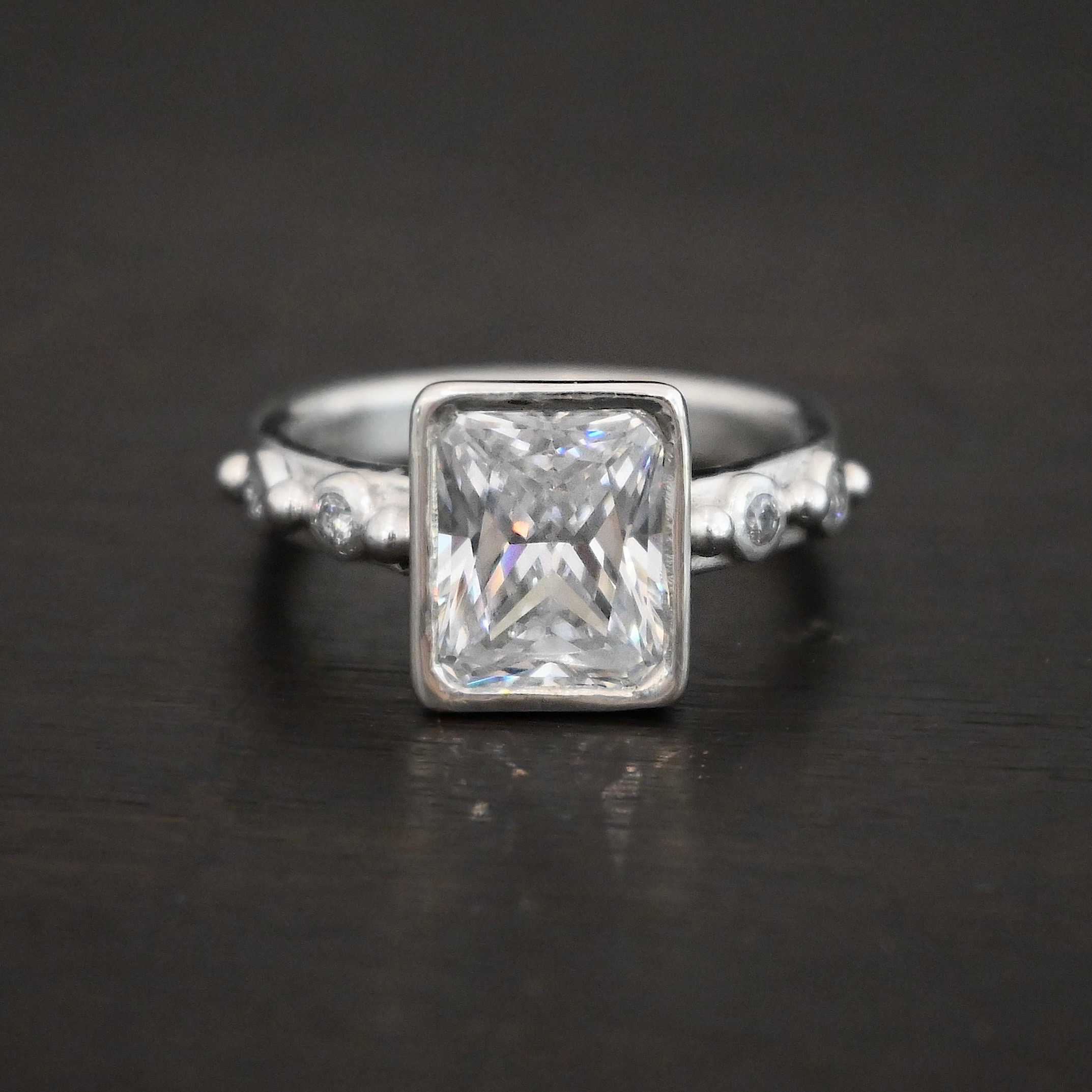 2 Have 2 Hold Ring Photo of the "2 Have and 2 Hold" Ring. There is a large rectangle moissanite in the center of the ring. On each side of the ring are two smaller moissanites, making four total. There are three silver balls separating the moissanites. One before the first moissanite, one in the center, and one on the end of the second moissanite. Ring is sitting against a dark background.