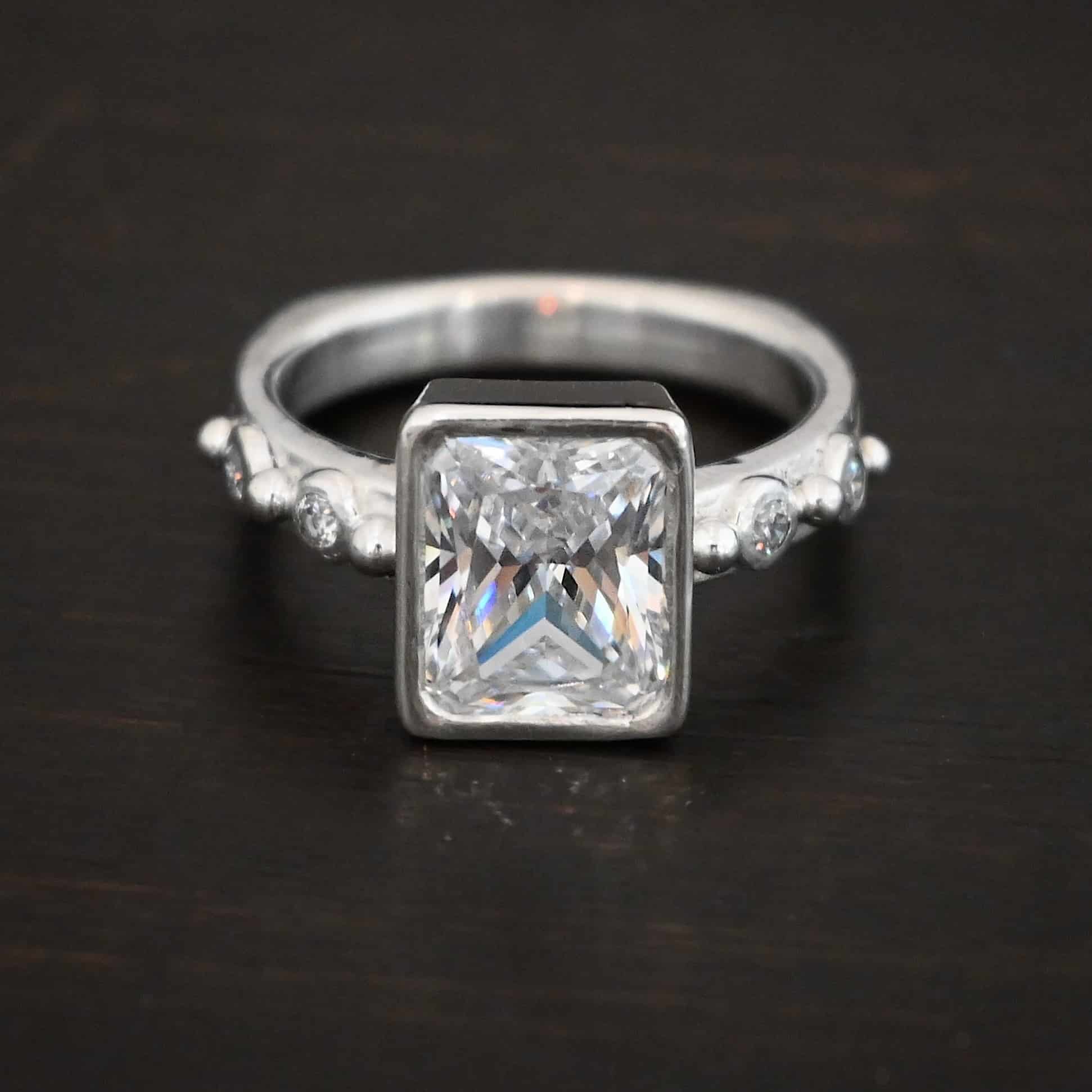 2 Have 2 Hold Ring Photo of the "2 Have and 2 Hold" Ring. There is a large rectangle moissanite in the center of the ring. On each side of the ring are two smaller moissanites, making four total. There are three silver balls separating the moissanites. One before the first moissanite, one in the center, and one on the end of the second moissanite. Ring is sitting against a dark background.