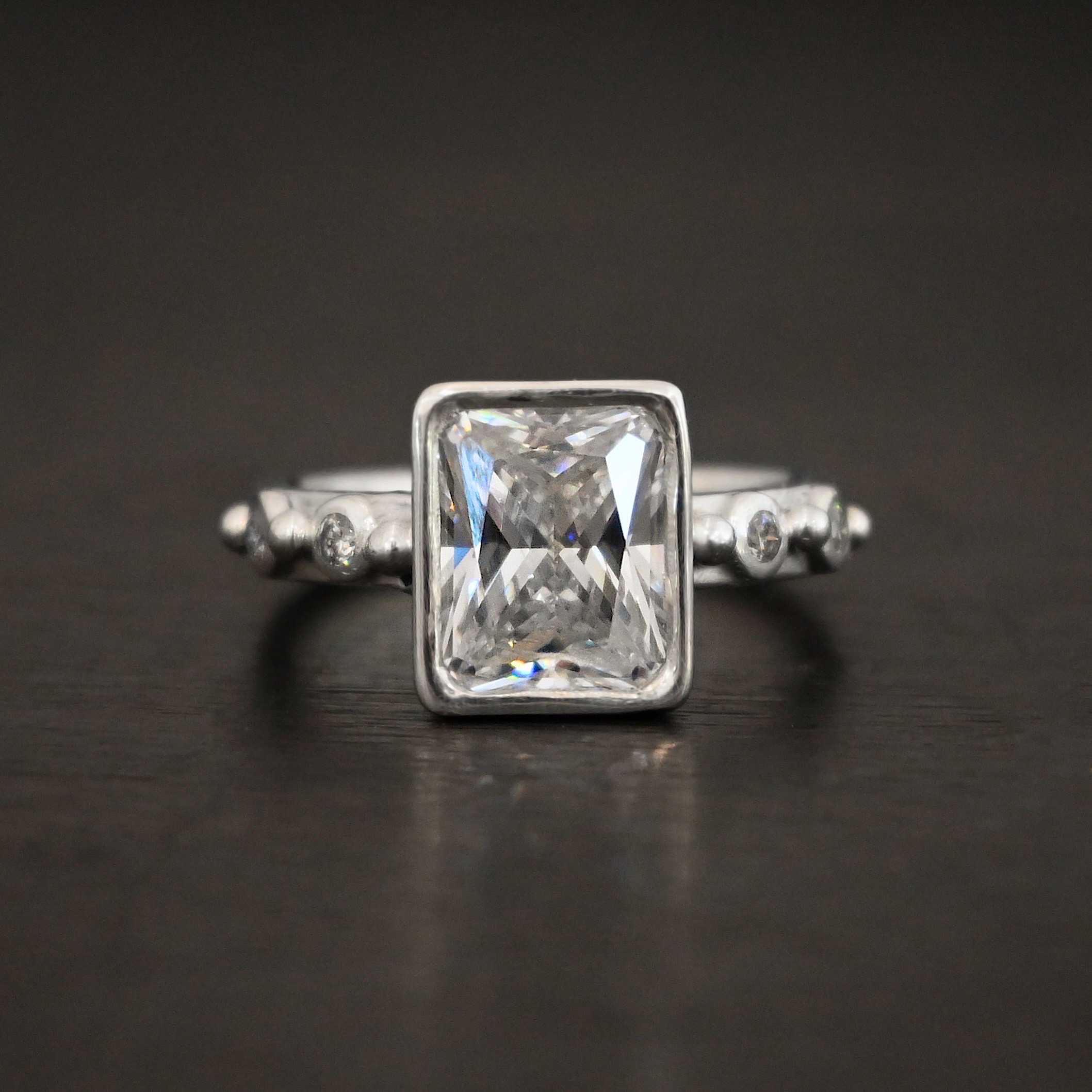 2 Have 2 Hold Ring Photo of the "2 Have and 2 Hold" Ring. There is a large rectangle moissanite in the center of the ring. On each side of the ring are two smaller moissanites, making four total. There are three silver balls separating the moissanites. One before the first moissanite, one in the center, and one on the end of the second moissanite. Ring is sitting against a dark background.
