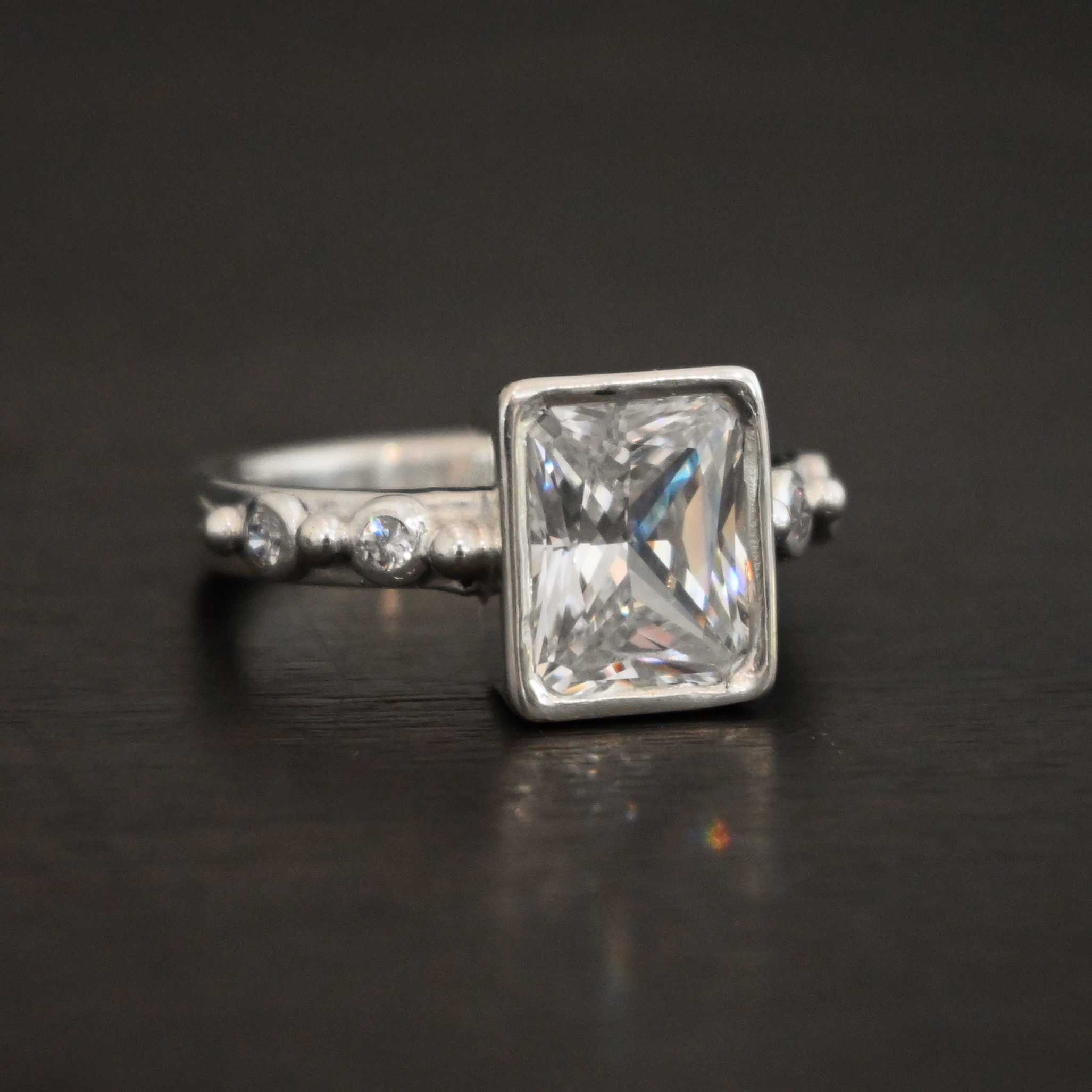 2 Have 2 Hold Ring Photo of the "2 Have and 2 Hold" Ring. There is a large rectangle moissanite in the center of the ring. On each side of the ring are two smaller moissanites, making four total. There are three silver balls separating the moissanites. One before the first moissanite, one in the center, and one on the end of the second moissanite. Ring is sitting against a dark background.