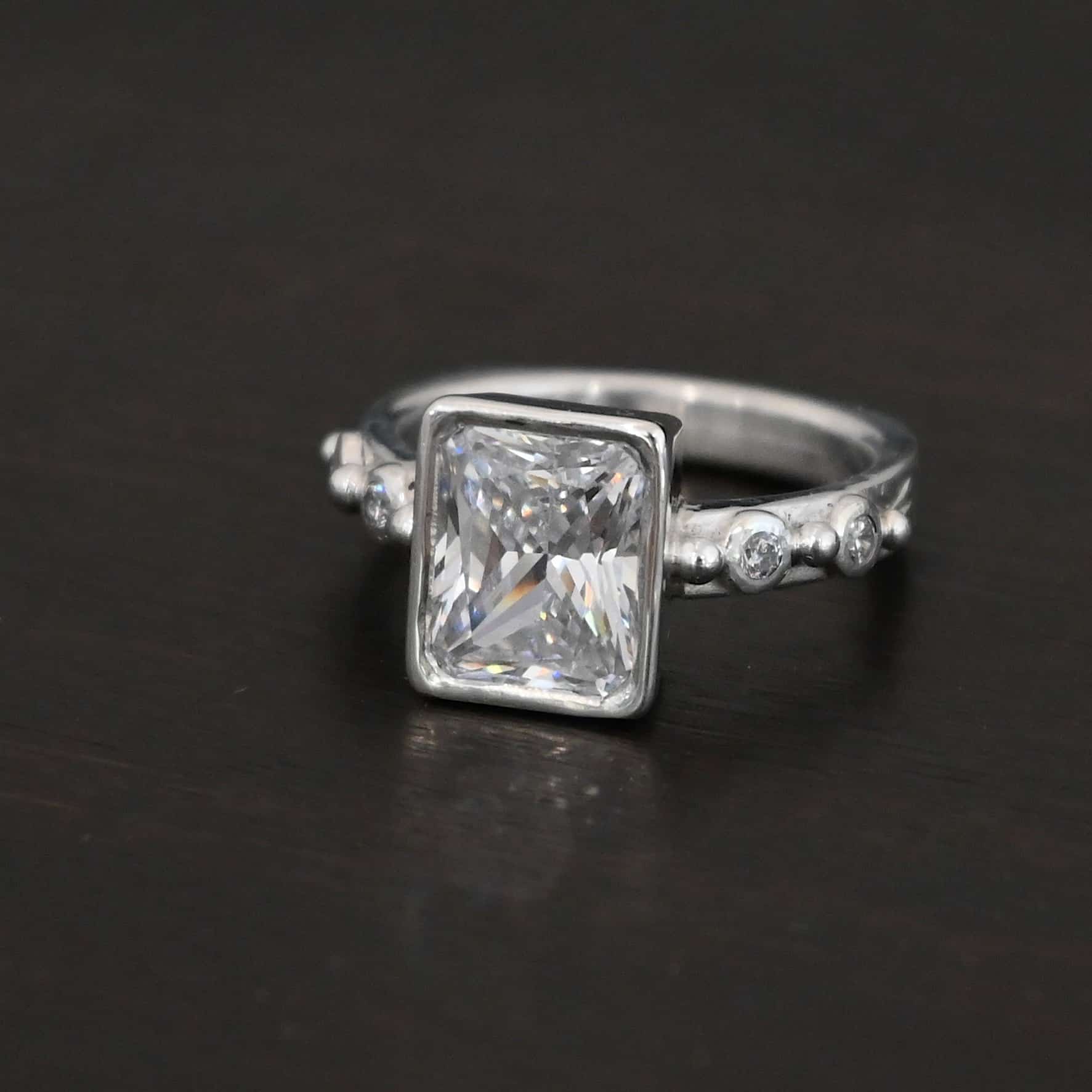 2 Have 2 Hold Ring Photo of the "2 Have and 2 Hold" Ring. There is a large rectangle moissanite in the center of the ring. On each side of the ring are two smaller moissanites, making four total. There are three silver balls separating the moissanites. One before the first moissanite, one in the center, and one on the end of the second moissanite. Ring is sitting against a dark background.