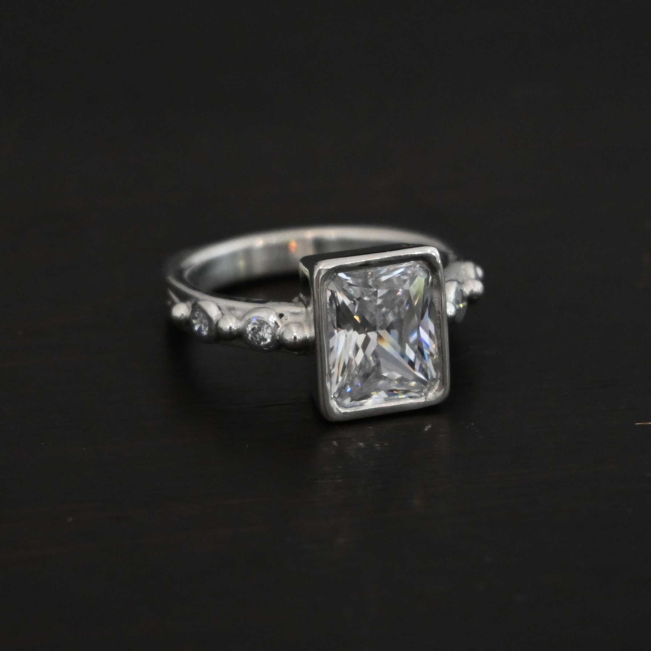 2 Have 2 Hold Ring Photo of the "2 Have and 2 Hold" Ring. There is a large rectangle moissanite in the center of the ring. On each side of the ring are two smaller moissanites, making four total. There are three silver balls separating the moissanites. One before the first moissanite, one in the center, and one on the end of the second moissanite. Ring is sitting against a dark background.