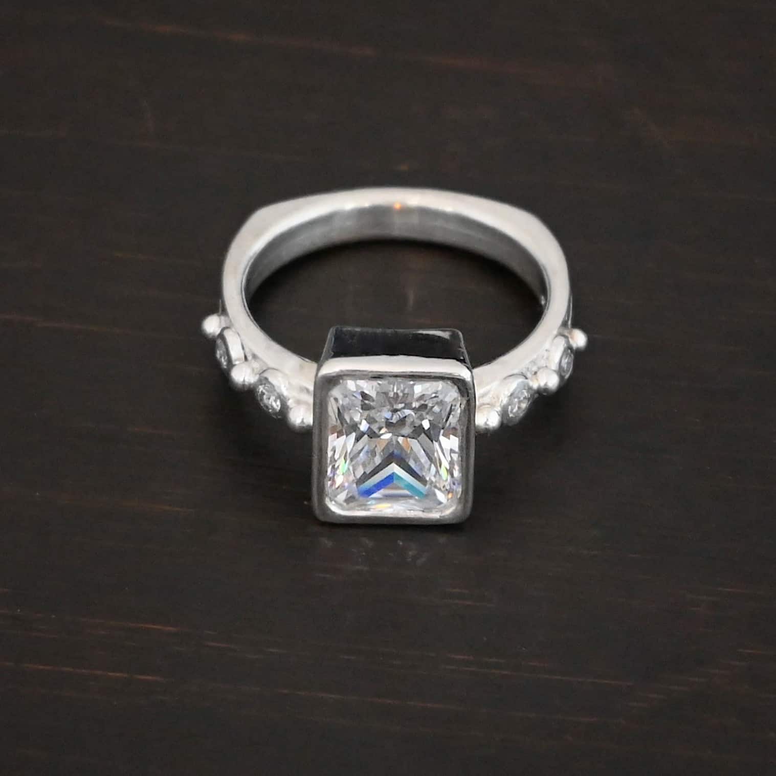2 Have 2 Hold Ring Photo of the "2 Have and 2 Hold" Ring. There is a large rectangle moissanite in the center of the ring. On each side of the ring are two smaller moissanites, making four total. There are three silver balls separating the moissanites. One before the first moissanite, one in the center, and one on the end of the second moissanite. Ring is sitting against a dark background.