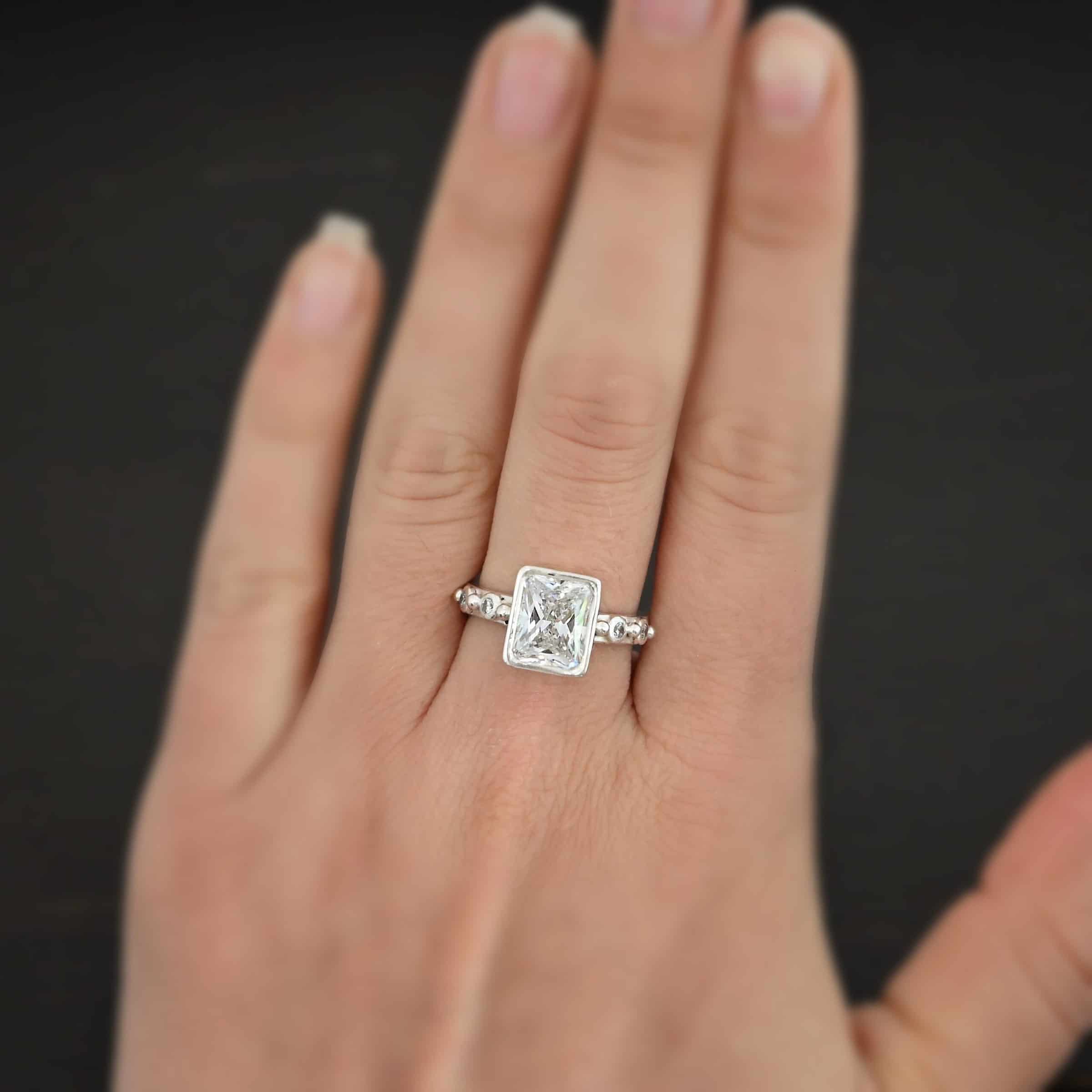 2 Have 2 Hold Ring Photo of the "2 Have and 2 Hold" Ring. There is a large rectangle moissanite in the center of the ring. On each side of the ring are two smaller moissanites, making four total. There are three silver balls separating the moissanites. One before the first moissanite, one in the center, and one on the end of the second moissanite. Ring is worn on a models engagement ring finger to demonstrate size.