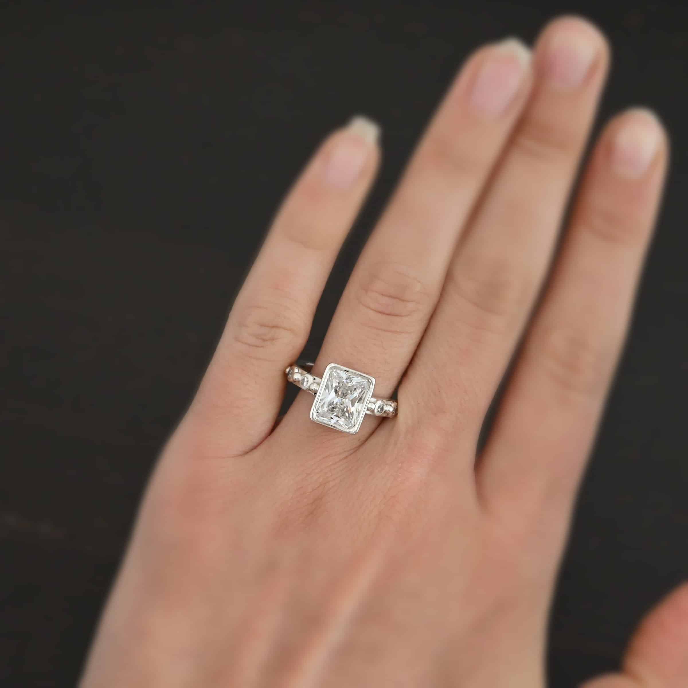 2 Have 2 Hold Ring Photo of the "2 Have and 2 Hold" Ring. There is a large rectangle moissanite in the center of the ring. On each side of the ring are two smaller moissanites, making four total. There are three silver balls separating the moissanites. One before the first moissanite, one in the center, and one on the end of the second moissanite. Ring is worn on a models engagement ring finger to demonstrate size.