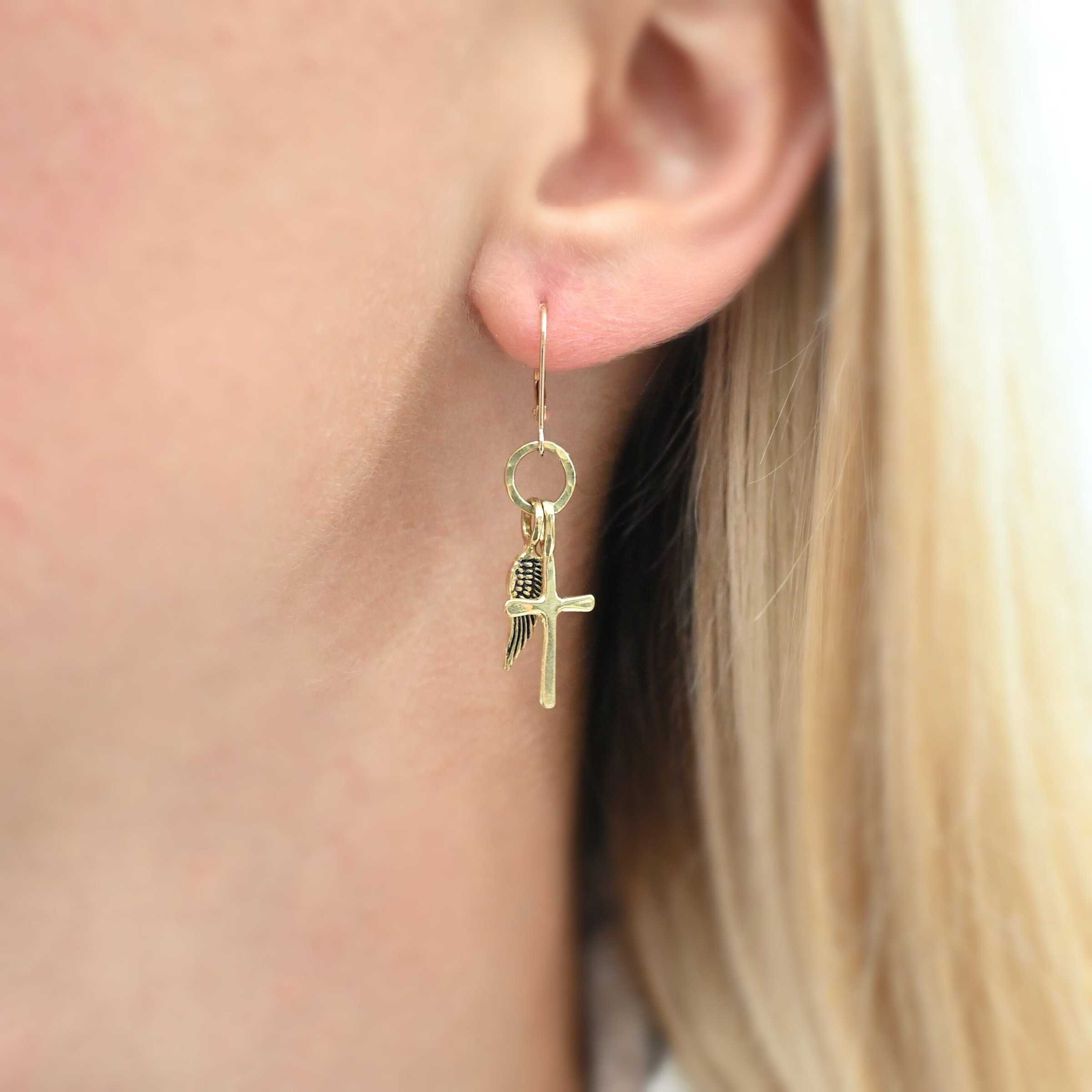 Strength Earrings 18k Photo of the "Strength" Earring as shown up close by a model. Made out of gold. There are a cross and angel wing charm. Both charms are small, and attached to a small hammered gold loop that the leverback ear wires are attached to on the top.