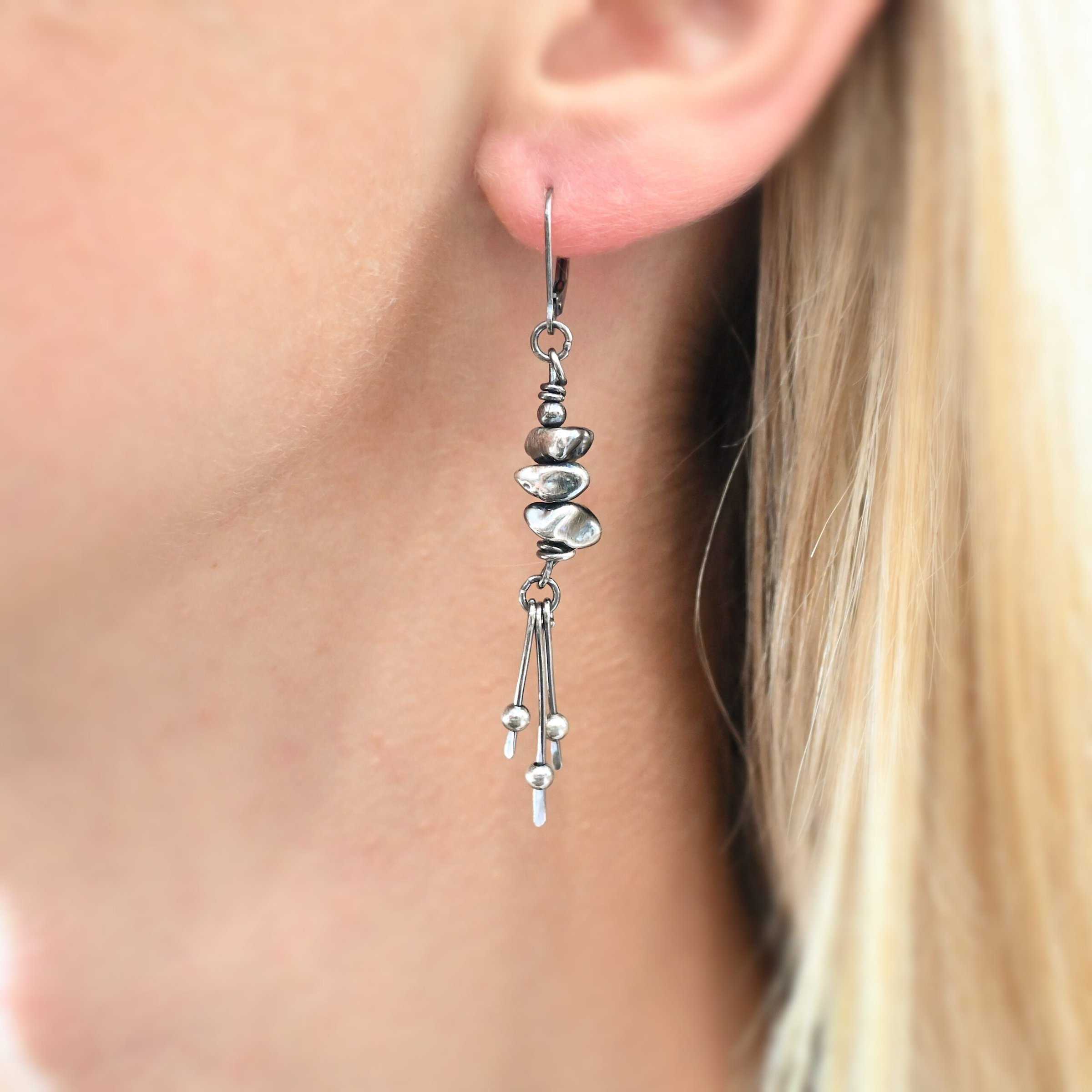 Photo of the "Pebble Beach" Earring as shown up close by a model. Made out of sterling silver. The earring has a leverback ear wire. The earring has three silver pebbles on top of one another beneath the ear wires. Hanging from the bottom of the pebbles are three short silver sticks with the ends hammered into paddles. Above the paddles are silver balls. There is one silver ball on each paddle.