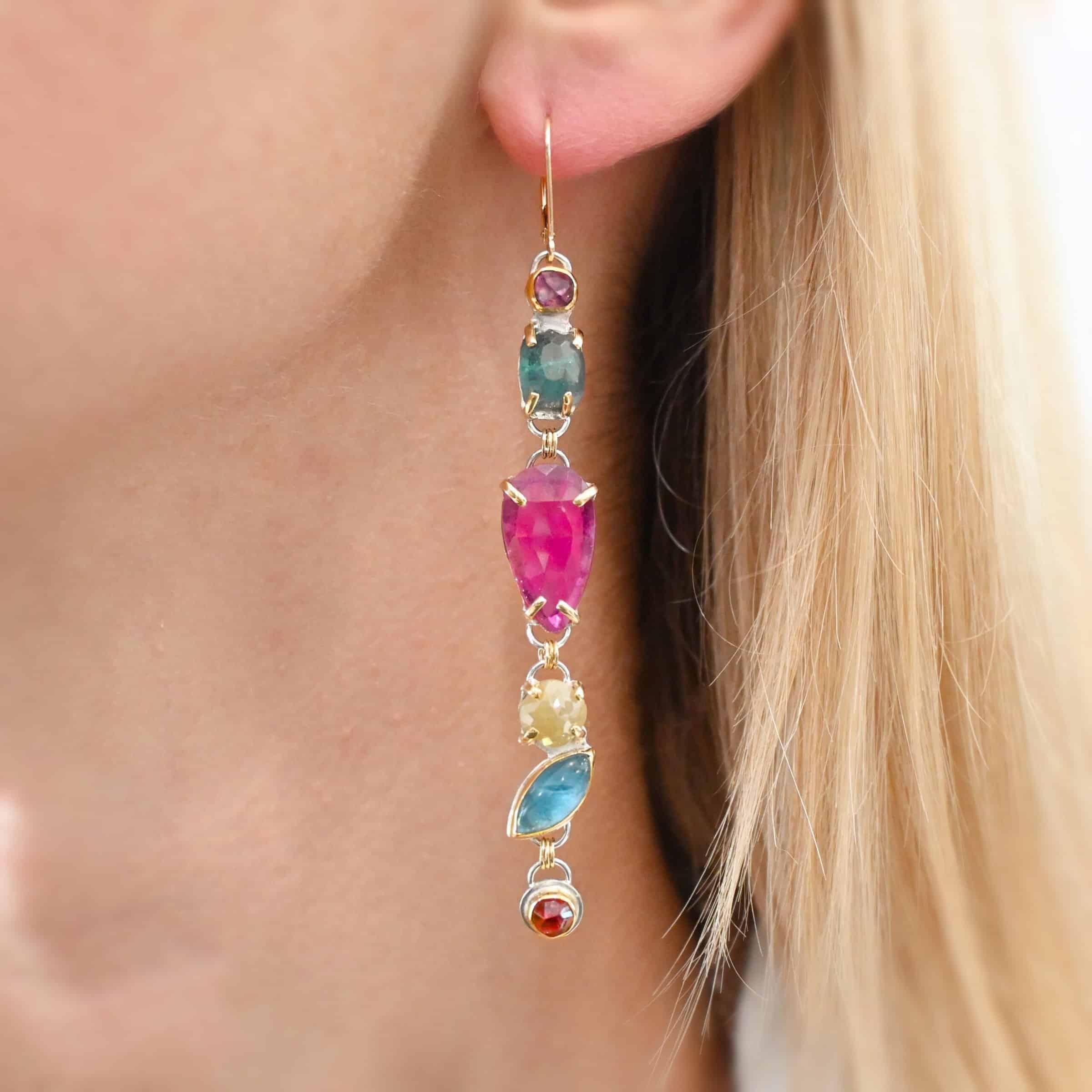 Follow Your Path Earrings Photo of the "Follow Your Path" Earring shown on a model up close. Made out of sterling silver & 18K gold, tourmalines, apatites, rough diamonds, & rubies. It is approximately 3.5″ long. The rubies are in the center, and the rest of the stones are above or beneath them. The rubies are held in with gold prongs, as well as some of the other stones. Each stone setting has a loop on the back of it with gold jump rings that connect the stones to one another. Earring has gold leverback ear wire.