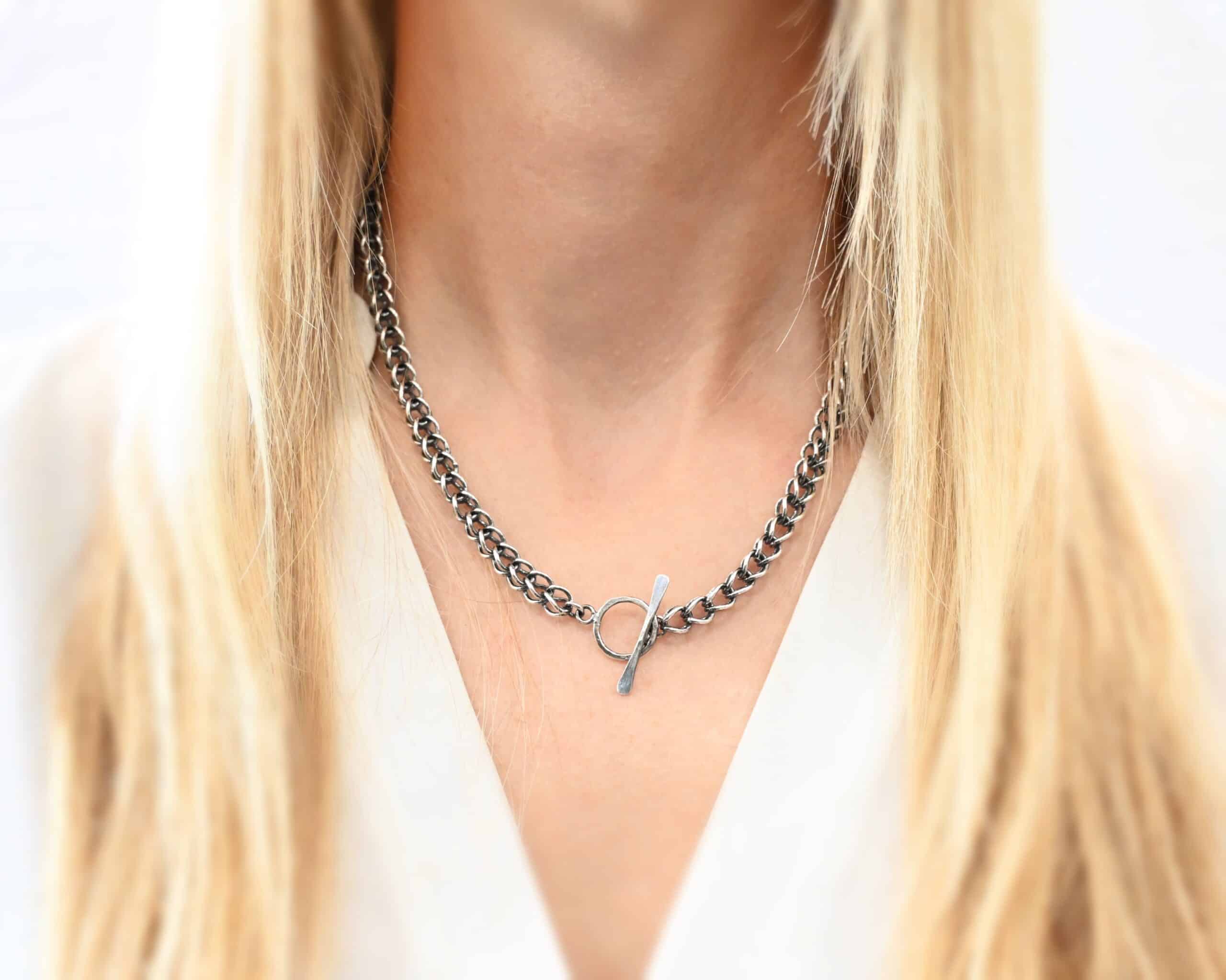 Gretchen Chain Photo of the "Gretchen" Chain. Chain is made out of sterling silver and oxidized. Chain is a viking knit chain, and approximately 18" long. Chain has a toggle clasp. Chain is flipped around to show the clasp of the necklace. Necklace is worn by a model. Model is wearing a white v-neck shirt.