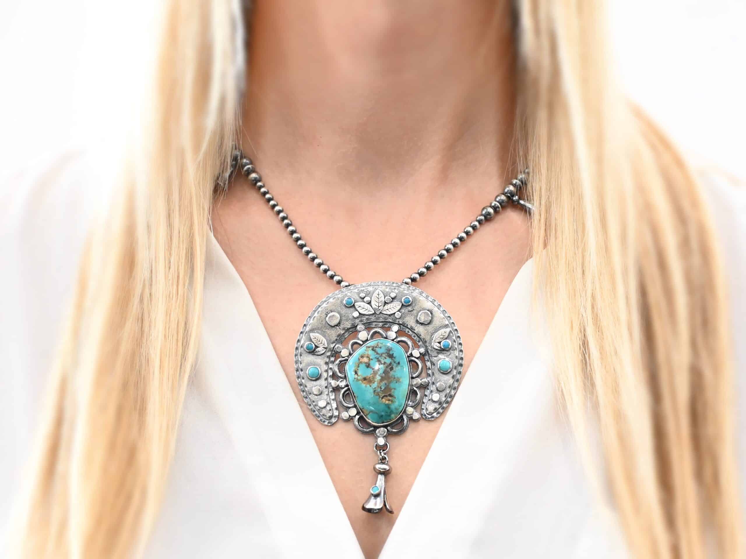 Photo of the "Dreamer Squash Blossom" Necklace. It has a large turquoise necklace. It is a squash blossom necklace. There is a large turquoise in the center with a flower petal design around it. There are seven other small turquoise on the horseshoe shaped silver piece going around the flower design of the turquoise. This piece has silver leaves and hammered silver dots on it as well. Hanging beneath the turquoise is a small cone of silver with cuts in it to make it look like an upside down flower petal. The chain is beaded and has the cone silver flower petals scattered around the chain. Necklace is worn by a model to demonstrate size.