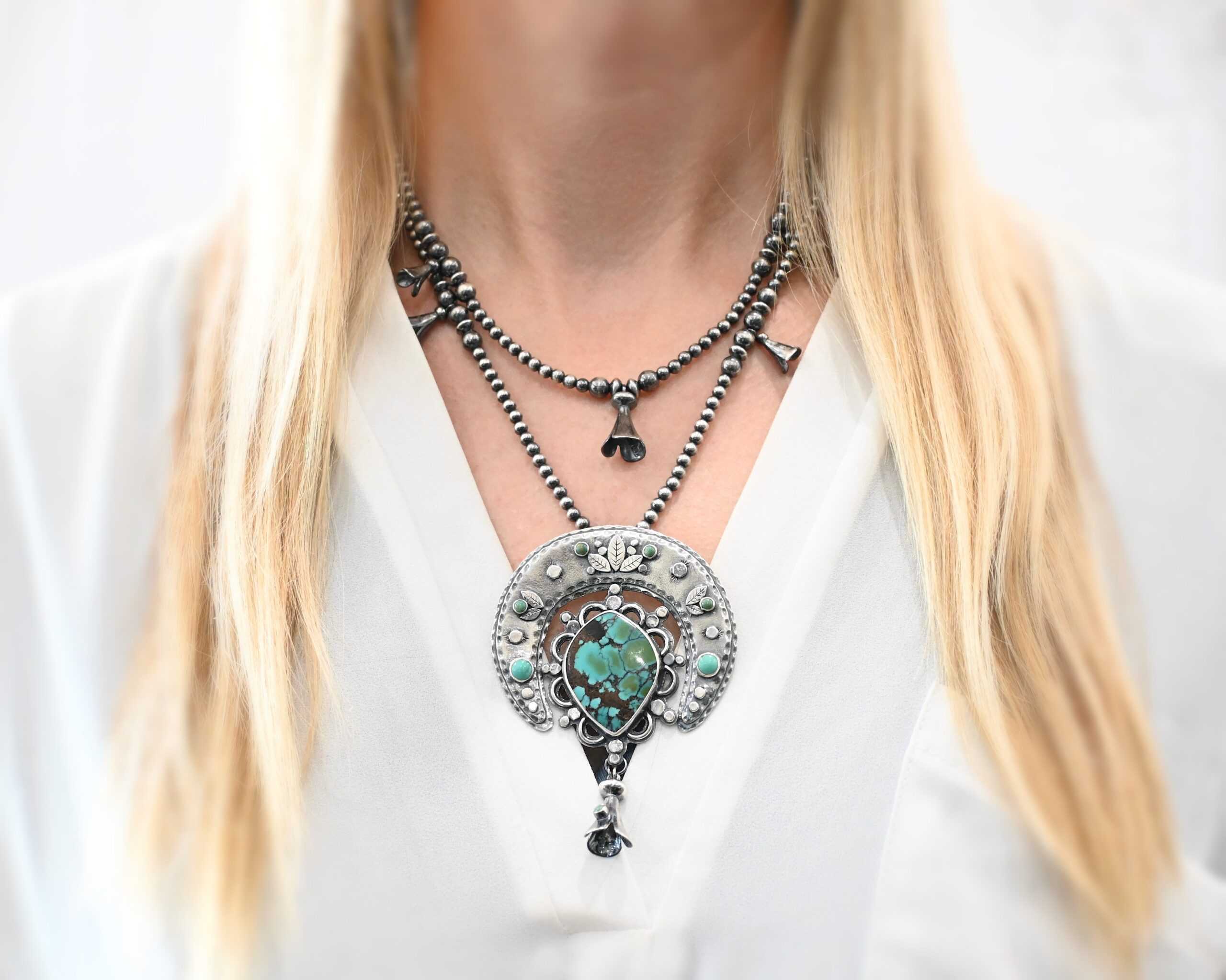 Wanderer Squash Blossom Necklace Photo of the "Wanderer Squash Blossom" Necklace. It has a large turquoise necklace. It is a squash blossom necklace. There is a large turquoise in the center with a flower petal design around it. There are seven other small turquoise on the horseshoe shaped silver piece going around the flower design of the turquoise. This piece has silver leaves and hammered silver dots on it as well. Hanging beneath the turquoise is a small cone of silver with cuts in it to make it look like an upside down flower petal. The chain is beaded and has the cone silver flower petals scattered around the chain. There is another beaded chain with cone flower petals on top of the necklace that goes with the necklace. Necklace is worn by a model to demonstrate size.