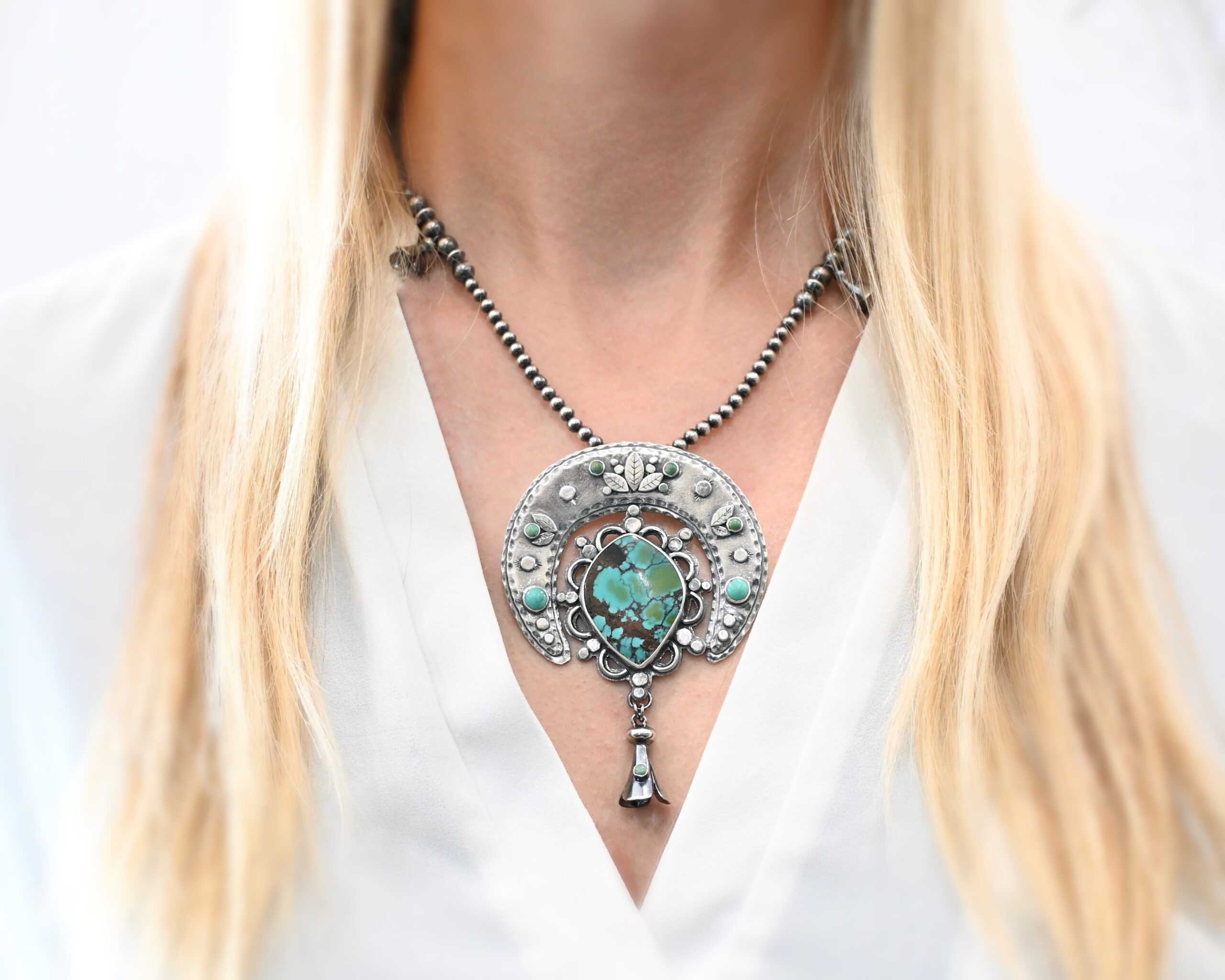 Wanderer Squash Blossom Necklace Photo of the "Wanderer Squash Blossom" Necklace. It has a large turquoise necklace. It is a squash blossom necklace. There is a large turquoise in the center with a flower petal design around it. There are seven other small turquoise on the horseshoe shaped silver piece going around the flower design of the turquoise. This piece has silver leaves and hammered silver dots on it as well. Hanging beneath the turquoise is a small cone of silver with cuts in it to make it look like an upside down flower petal. The chain is beaded and has the cone silver flower petals scattered around the chain. Necklace is worn by a model to demonstrate size.