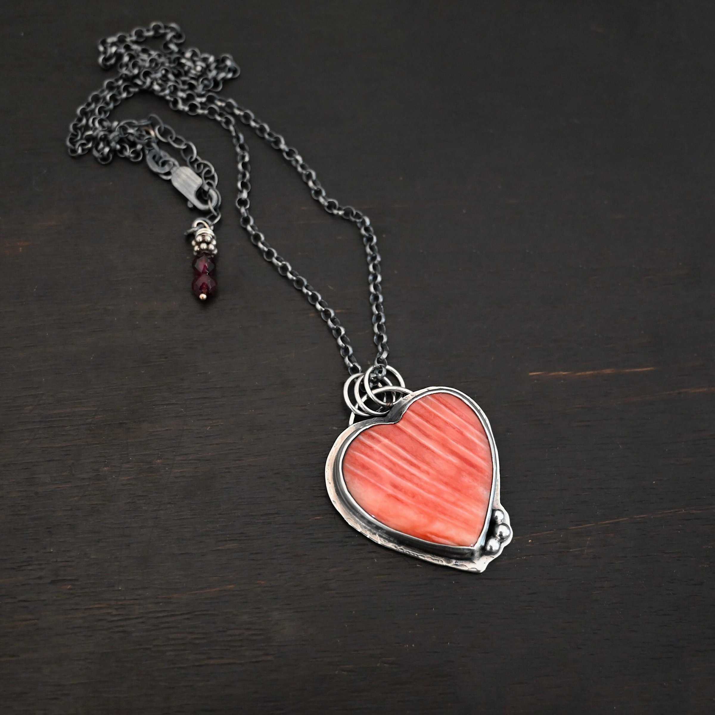 Photo of "A Weathered Heart" Necklace. Made out of sterling silver and spiny oyster. The pendant is a heart shaped spiny oyster cabochon. It is a red/orange color mix with some white lines going through it. There are three small silver balls on the bottom right side of the heart pendant. The two outermost balls are smaller, and the ball in the center is larger. They are placed right next to one another, and are close to the tip of the heart. There is a loop on the back of the pendant with three jump rings attached to it. Going through the jump rings are the chain of the necklace. On the end of the extension for the necklace are two garnet beads. Necklace is laying against a dark background.