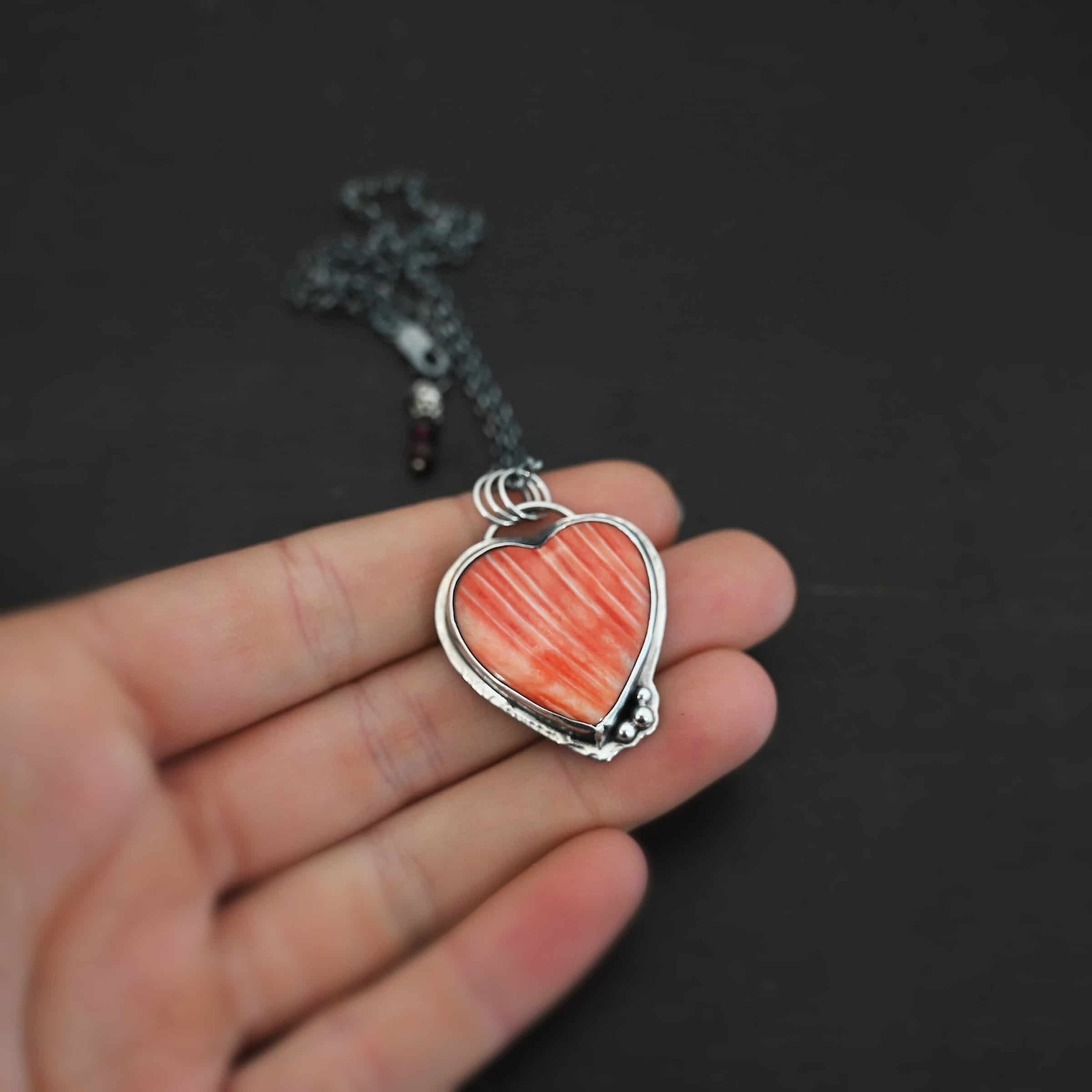 Photo of "A Weathered Heart" Necklace. Made out of sterling silver and spiny oyster. The pendant is a heart shaped spiny oyster cabochon. It is a red/orange color mix with some white lines going through it. There are three small silver balls on the bottom right side of the heart pendant. The two outermost balls are smaller, and the ball in the center is larger. They are placed right next to one another, and are close to the tip of the heart. There is a loop on the back of the pendant with three jump rings attached to it. Going through the jump rings are the chain of the necklace. On the end of the extension for the necklace are two garnet beads. Necklace is laying against the fingertips of a hand to demonstrate size.
