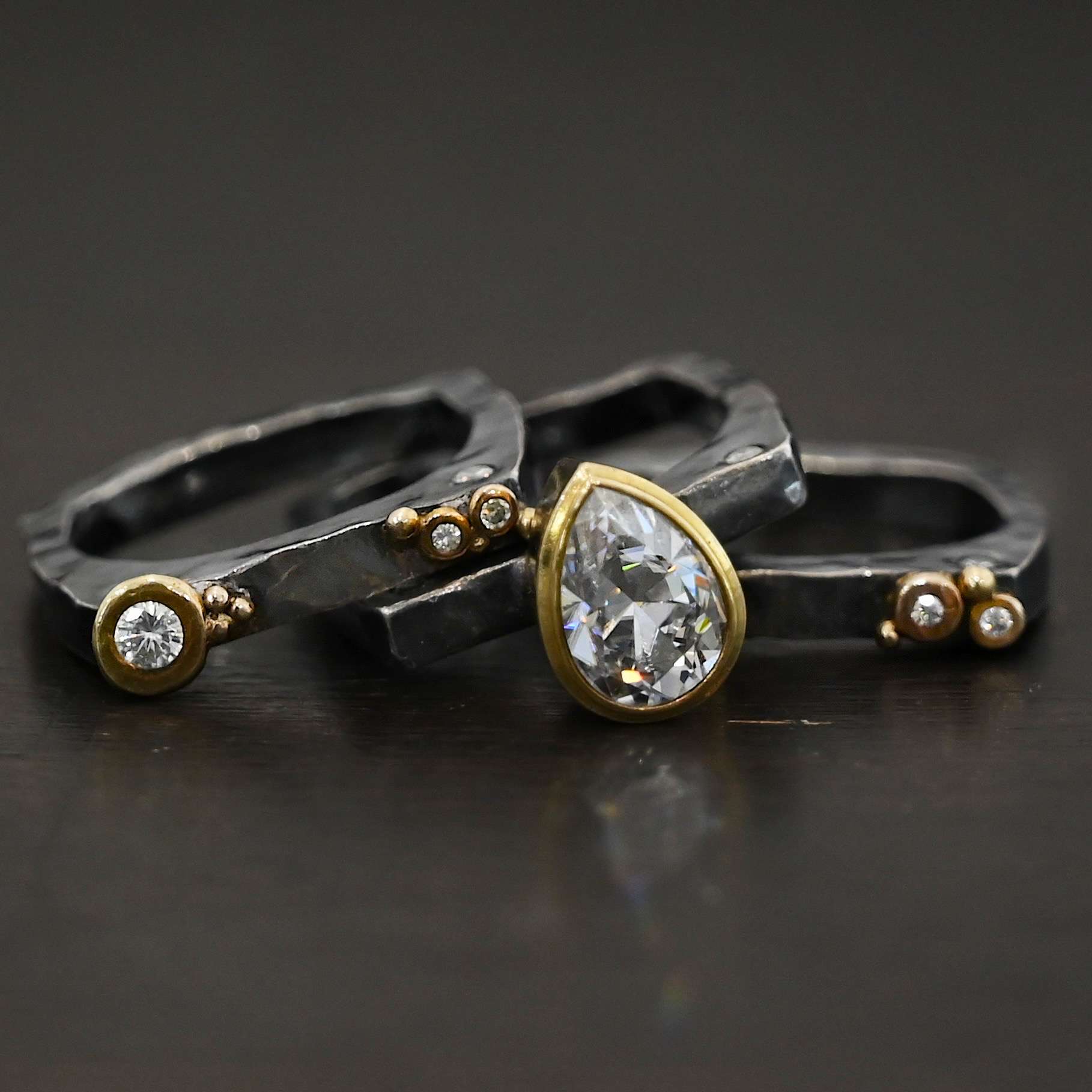 Photo of the Johnny & June Ring. Made out of sterling silver, gold, and moissanites. There is a pear shaped moissanite in the center. The ring is three separate stacker bands. There are three moissanites that are smaller on the top band and three on the bottom band. The large teardrop center stone spans across all three bands. There is gold ball detail across all three bands. Ring is sitting against a dark background, with all three bands separated and laying on top of one another.