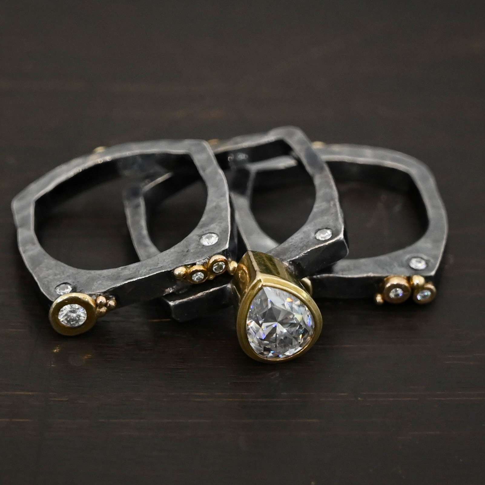 Photo of the Johnny & June Ring. Made out of sterling silver, gold, and moissanites. There is a pear shaped moissanite in the center. The ring is three separate stacker bands. There are three moissanites that are smaller on the top band and three on the bottom band. The large teardrop center stone spans across all three bands. There is gold ball detail across all three bands. Ring is sitting against a dark background, with all three bands separated and laying on top of one another.