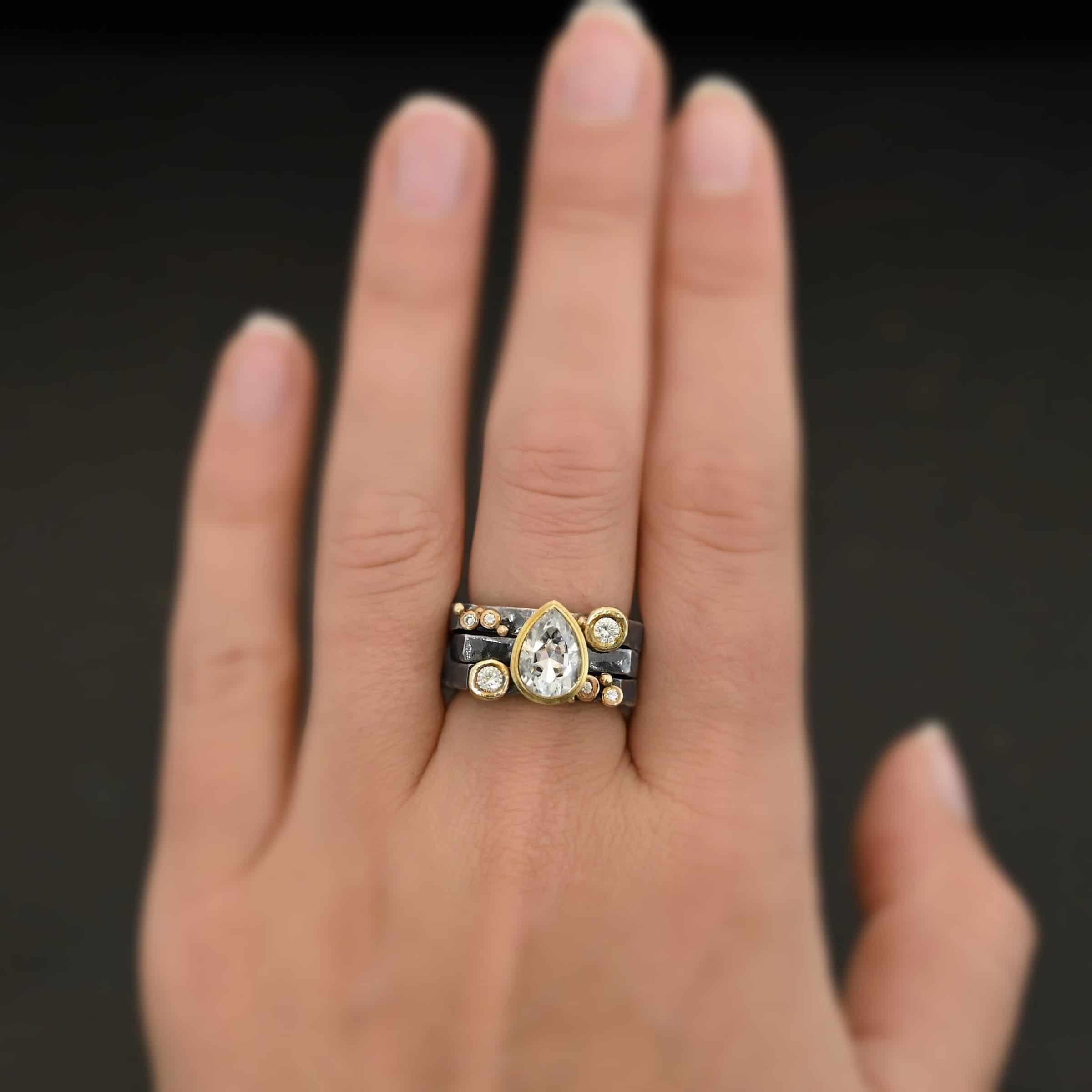 Photo of the Johnny & June Ring. Made out of sterling silver, gold, and moissanites. There is a pear shaped moissanite in the center. The ring is three separate stacker bands. There are three moissanites that are smaller on the top band and three on the bottom band. The large teardrop center stone spans across all three bands. There is gold ball detail across all three bands. Ring is worn on a models middle finger to demonstrate size.