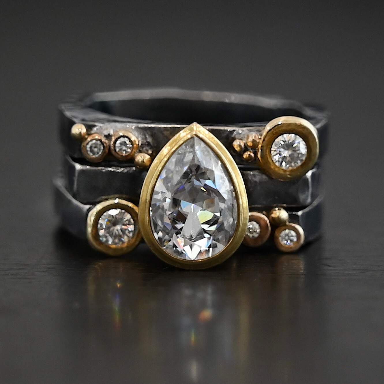 Photo of the Johnny & June Ring. Made out of sterling silver, gold, and moissanites. There is a pear shaped moissanite in the center. The ring is three separate stacker bands. There are three moissanites that are smaller on the top band and three on the bottom band. The large teardrop center stone spans across all three bands. There is gold ball detail across all three bands. Ring is sitting against a dark background.