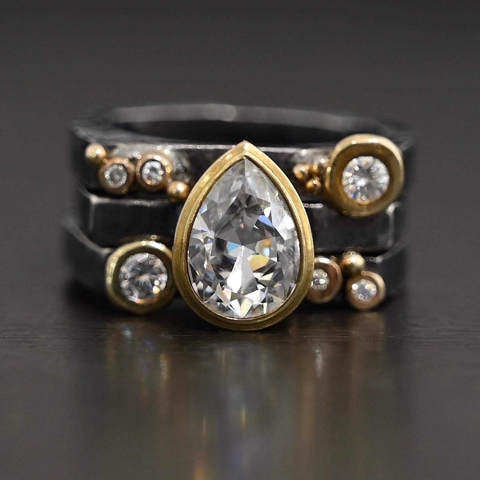 Photo of the Johnny & June Ring. Made out of sterling silver, gold, and moissanites. There is a pear shaped moissanite in the center. The ring is three separate stacker bands. There are three moissanites that are smaller on the top band and three on the bottom band. The large teardrop center stone spans across all three bands. There is gold ball detail across all three bands. Ring is sitting against a dark background.