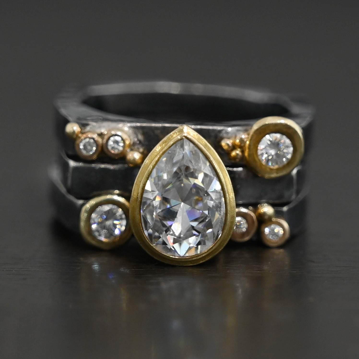 Photo of the Johnny & June Ring. Made out of sterling silver, gold, and moissanites. There is a pear shaped moissanite in the center. The ring is three separate stacker bands. There are three moissanites that are smaller on the top band and three on the bottom band. The large teardrop center stone spans across all three bands. There is gold ball detail across all three bands. Ring is sitting against a dark background.