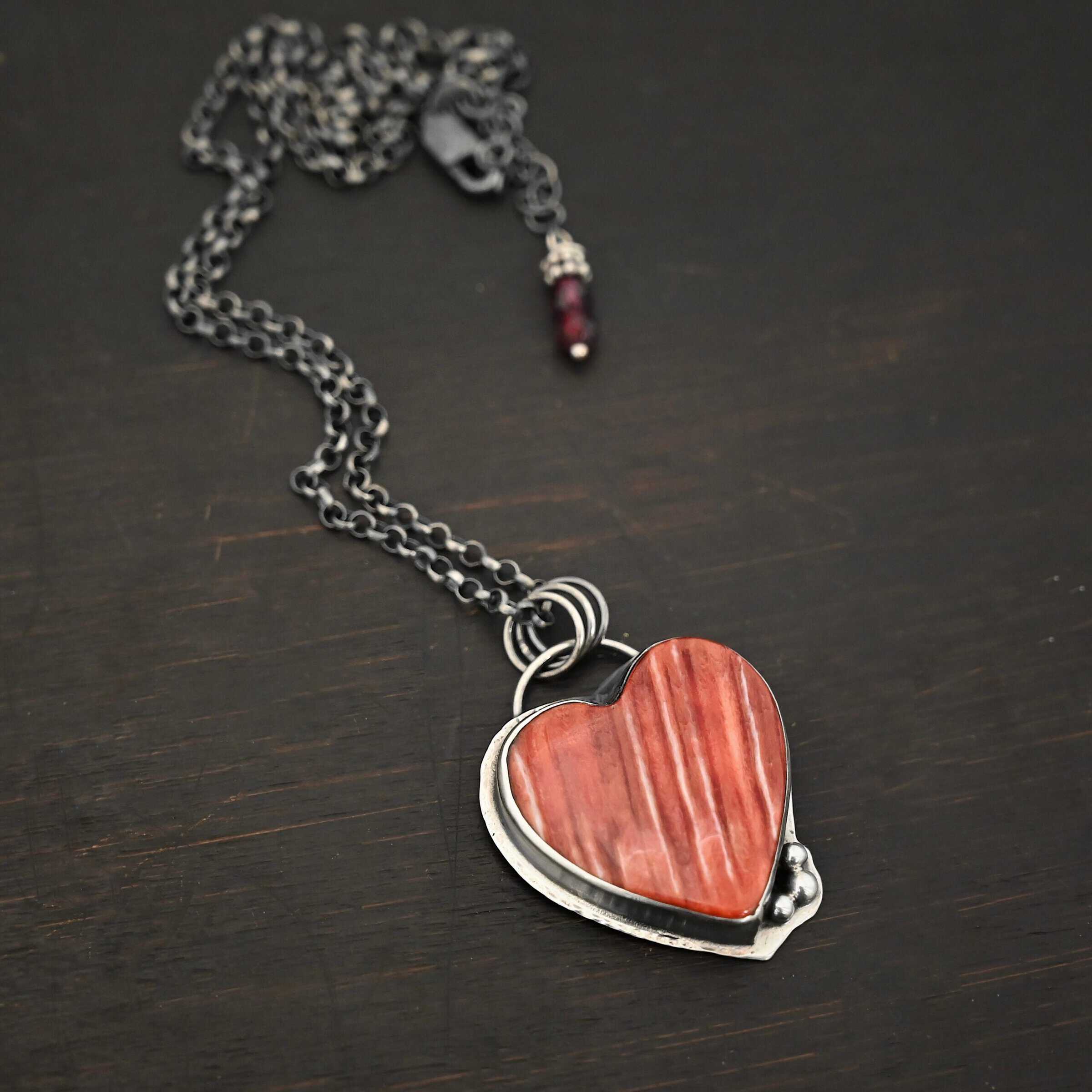 Photo of "A Weathered Heart" Necklace. Made out of sterling silver and spiny oyster. The pendant is a heart shaped spiny oyster cabochon. It is a red/orange color mix with some white lines going through it. There are three small silver balls on the bottom right side of the heart pendant. The two outermost balls are smaller, and the ball in the center is larger. They are placed right next to one another, and are close to the tip of the heart. There is a loop on the back of the pendant with three jump rings attached to it. Going through the jump rings are the chain of the necklace. On the end of the extension for the necklace are two garnet beads. Necklace is laying against a dark background.