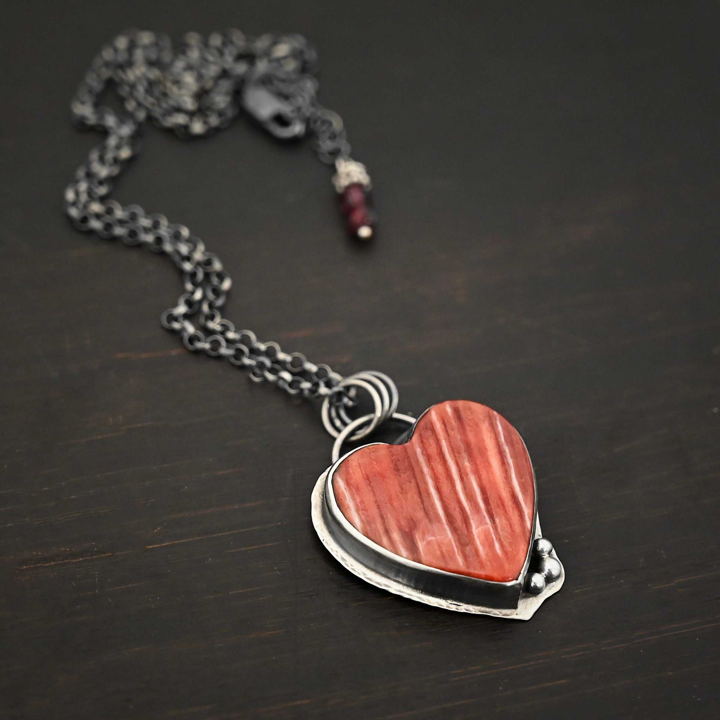 Photo of "A Weathered Heart" Necklace. Made out of sterling silver and spiny oyster. The pendant is a heart shaped spiny oyster cabochon. It is a red/orange color mix with some white lines going through it. There are three small silver balls on the bottom right side of the heart pendant. The two outermost balls are smaller, and the ball in the center is larger. They are placed right next to one another, and are close to the tip of the heart. There is a loop on the back of the pendant with three jump rings attached to it. Going through the jump rings are the chain of the necklace. On the end of the extension for the necklace are two garnet beads. Necklace is laying against a dark background.